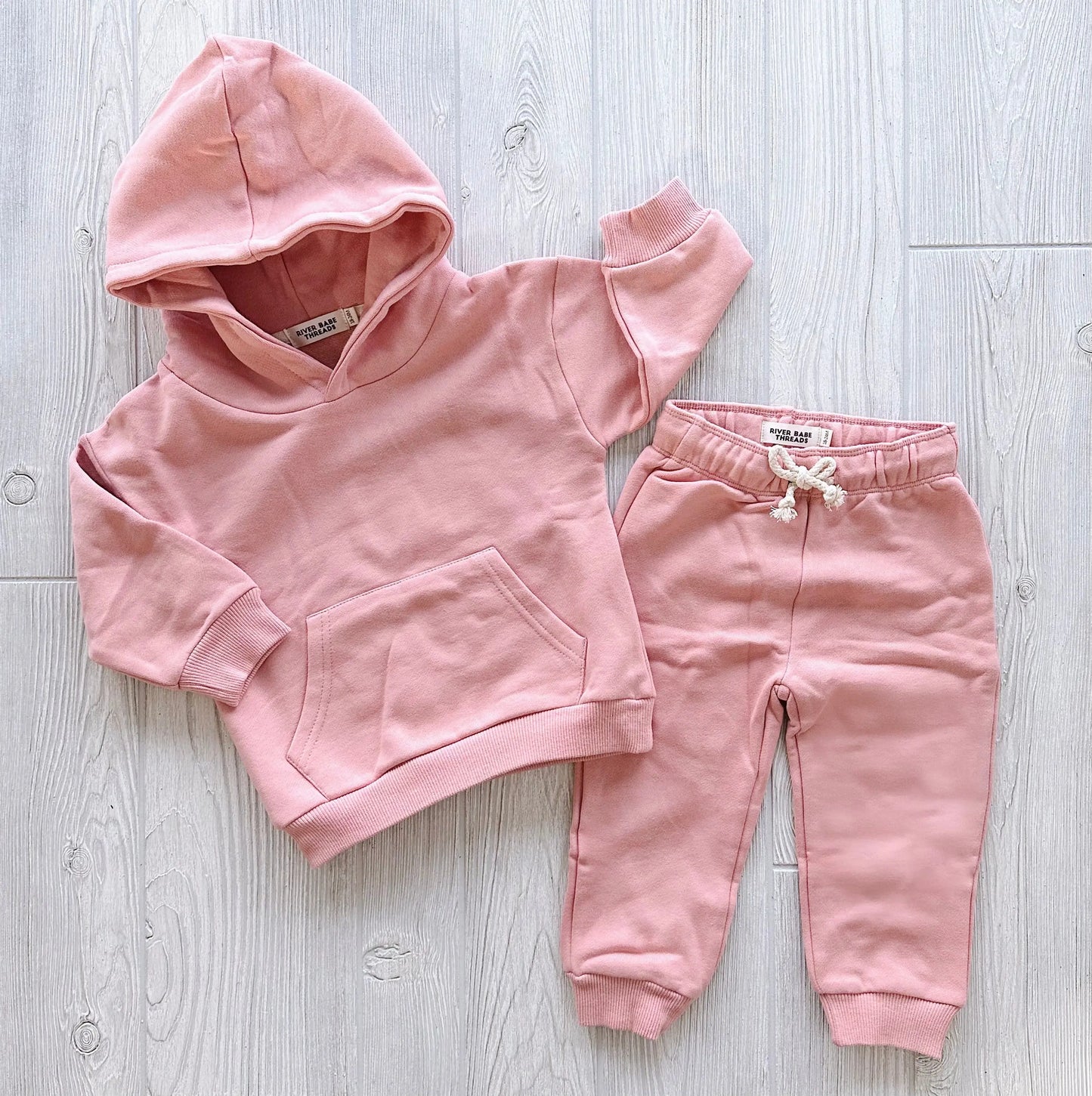 The Mauve • Sweatpant & Hoodie Set is an organic two-piece in pink, featuring a fleece hoodie with a front pocket and matching adjustable drawstring sweatpants, displayed on a light wooden floor.