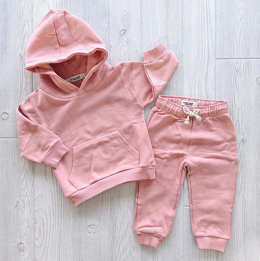 The Mauve • Sweatpant & Hoodie Set is an organic two-piece in pink, featuring a fleece hoodie with a front pocket and matching adjustable drawstring sweatpants, displayed on a light wooden floor.