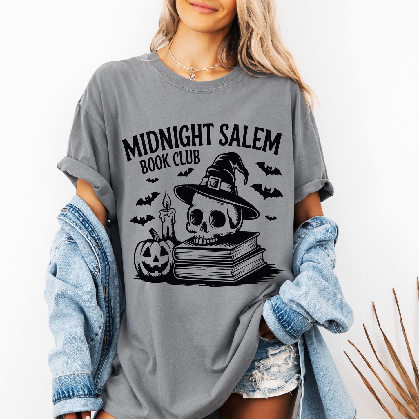 A woman wears the Midnight Salem Book Club • Comfort Tee in gray, showcasing a skull with witch hat, candle, bats, stacked books, and a jack-o-lantern. She pairs this garment-dyed shirt with a denim jacket and shorts for her zombie-inspired look.