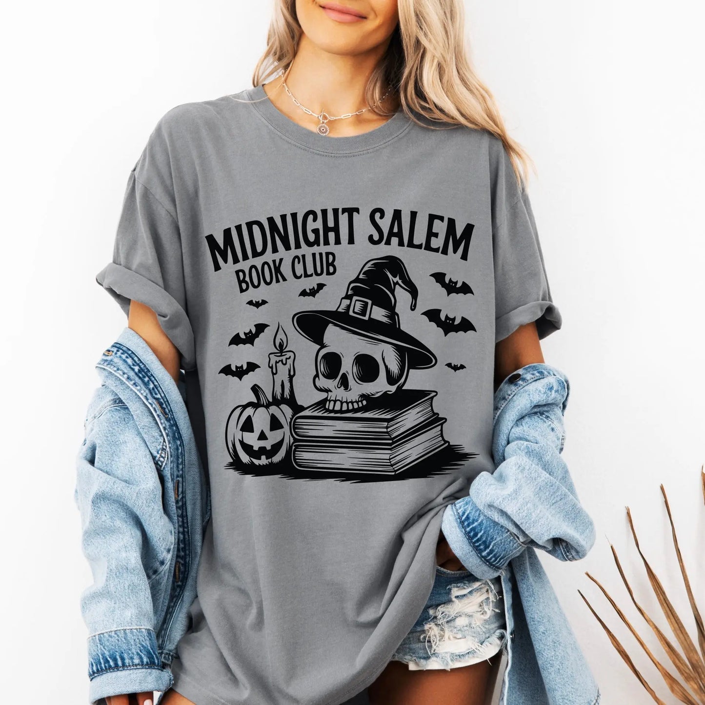 A woman wears the Midnight Salem Book Club • Comfort Tee in gray, showcasing a skull with witch hat, candle, bats, stacked books, and a jack-o-lantern. She pairs this garment-dyed shirt with a denim jacket and shorts for her zombie-inspired look.