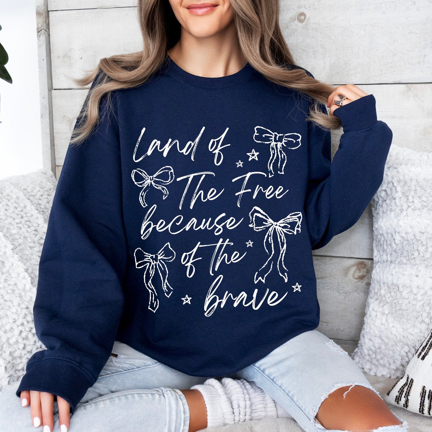 A woman’s lower face is visible as she sits on a couch wearing the Because of the Brave • Navy Pullover, a unisex sweatshirt featuring white “Land of The Free because of the brave” text with bows and stars.