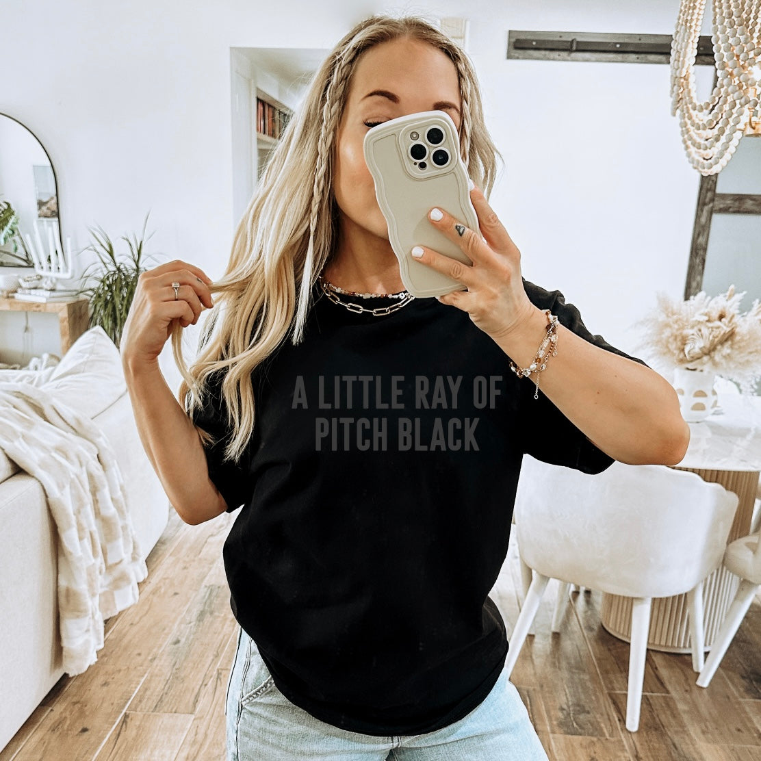 A woman with long blonde hair takes a mirror selfie in a stylish living room, wearing the Pitch Black • Tonal Tee with A LITTLE RAY OF PITCH BLACK text. She holds her phone up, hiding her face, and pairs it with light blue jeans.