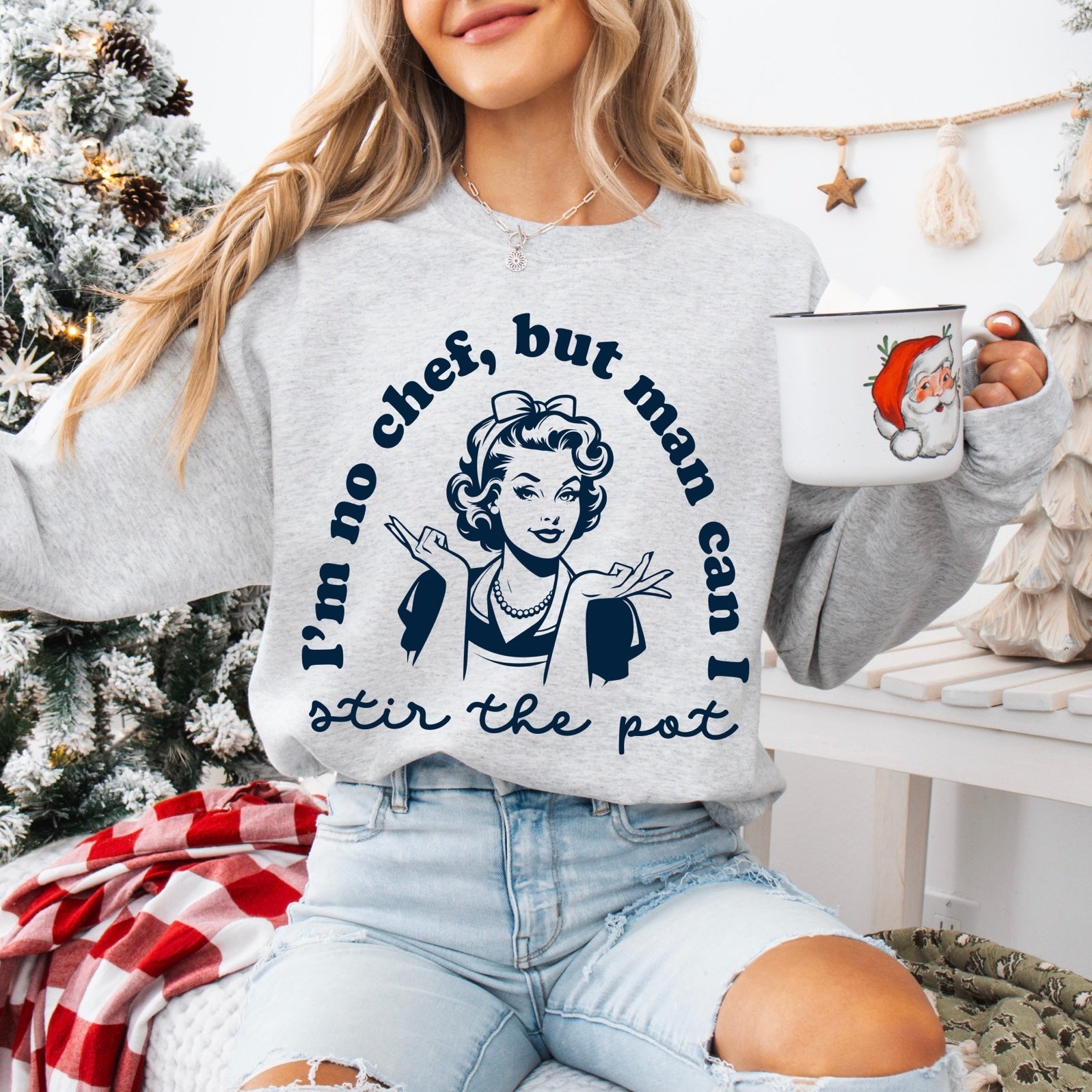 Wearing the Stir the Pot • Ash Pullover, a woman with a retro “Im no chef, but man can I stir the pot” design relaxes by a decorated Christmas tree, smiling and holding a festive mug.