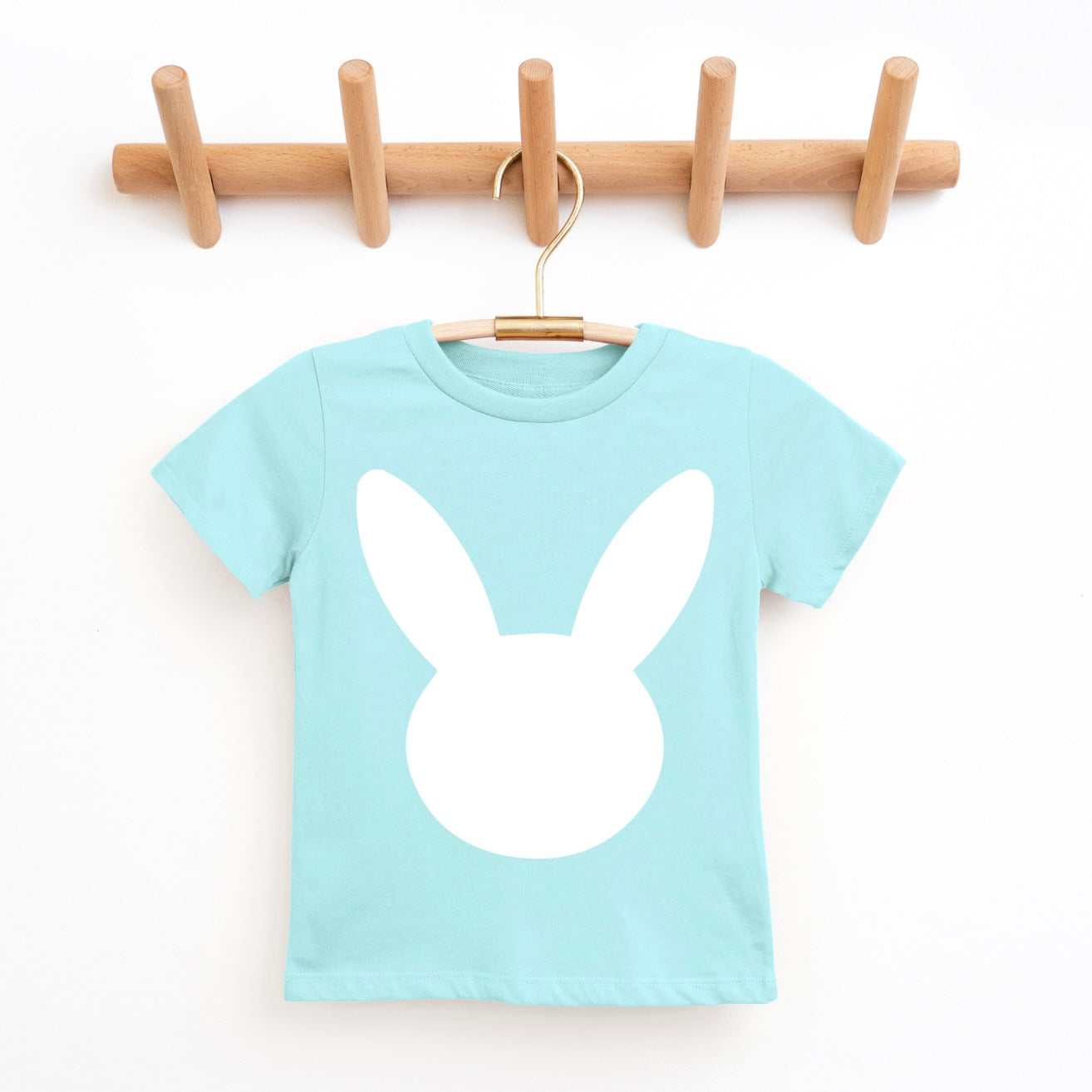 Introducing the NEW! Bunny • Pastel Teal Tee—a unisex childrens t-shirt in light blue, featuring a large white Bunny design on the front, displayed on a wooden hanger from a wall-mounted peg rack.