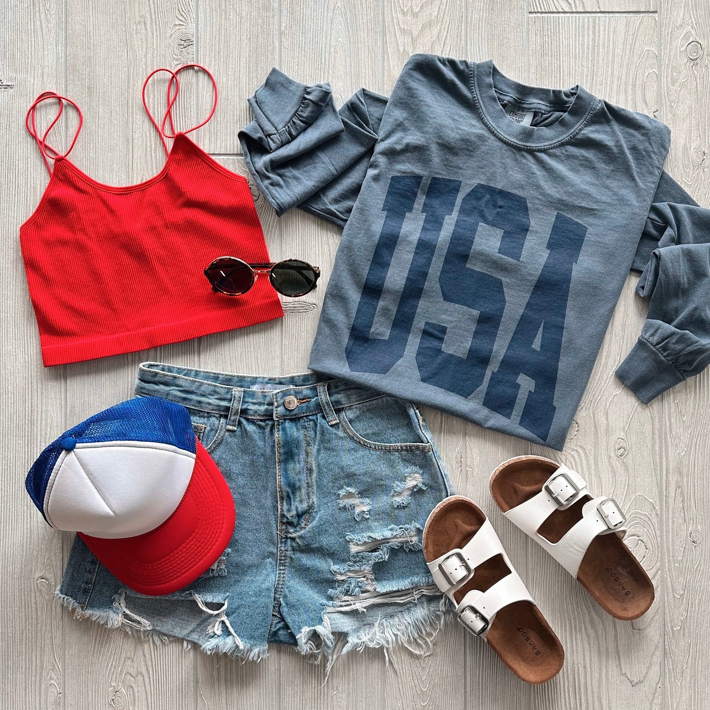 Flat lay of a casual summer outfit with a red crop top, denim shorts, white slide sandals, round black sunglasses, a red, white, and blue mesh cap, and the USA • Tonal Navy Long Sleeve on light wood floor.