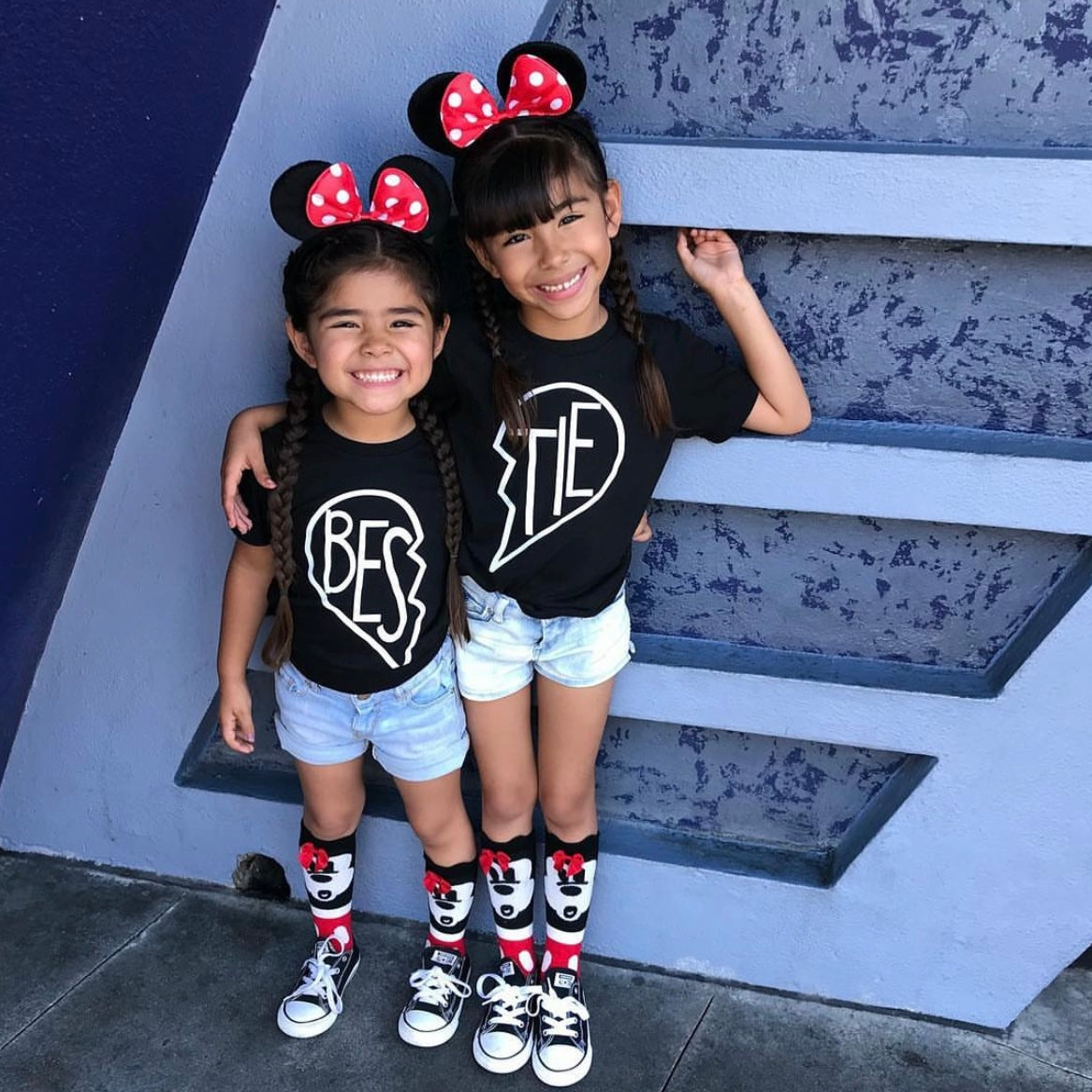Two young girls with braided hair smile and hug wearing matching unisex Bestie • Black Tees, Minnie Mouse ears, denim shorts, and Minnie Mouse socks in front of a blue textured wall.