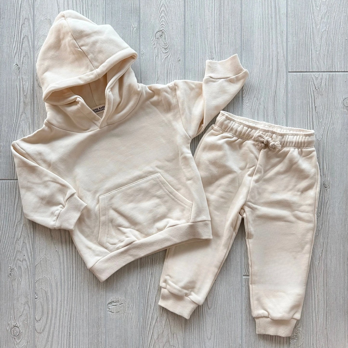 The Oatmeal • Sweatpant & Hoodie Set, an organic cream two-piece with a fleece hoodie (front pocket) and matching drawstring sweatpants, is displayed laid flat on a light wood floor.