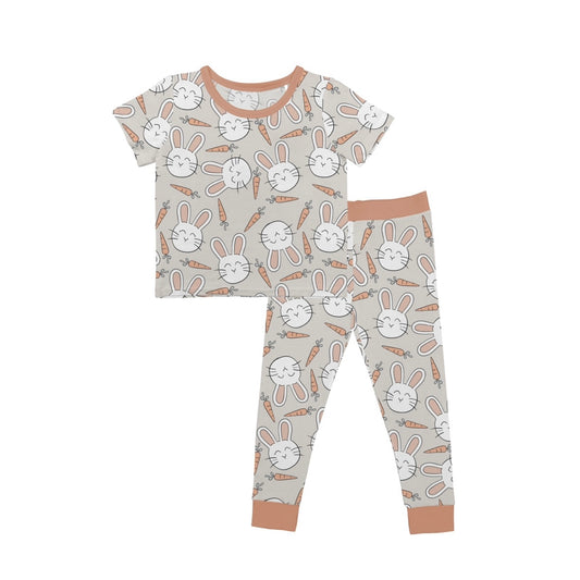 Introducing the Snuggle Bunny • 2 Piece Bamboo Set (Updated Fit): a cozy children’s pajama set with a beige background, smiling white bunnies, and orange carrots. Comes with a short-sleeve top and long pants trimmed in light brown, made from ultra-soft bamboo fabric.
