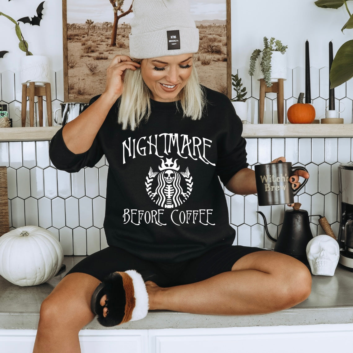 A smiling woman in a beanie and a Nightmare Before Coffee • Tee or Sweatshirt by Bella/Canvas sits on a kitchen counter holding a Witch’s Brew mug, surrounded by Halloween decorations, pumpkins, and plants.