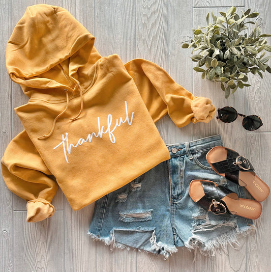 Flat lay of the "thankful • Mustard Hoodie" styled with distressed denim shorts, black sandals, round sunglasses, and a small potted plant—showcasing perfect autumn hues on a light wood floor.