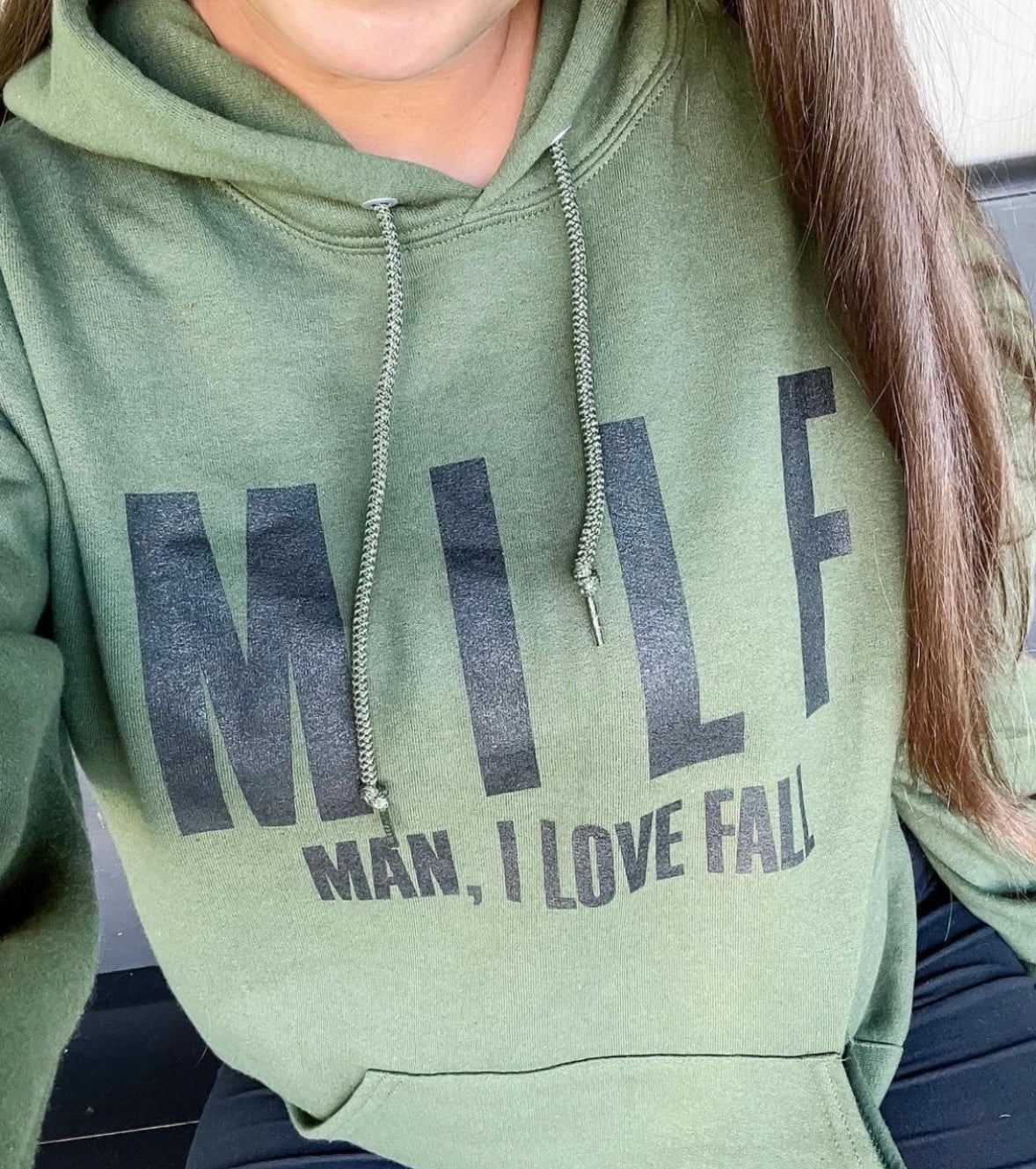 A person wearing the unisex MILF • Olive Hoodie, featuring large black MILF letters and smaller Man, I Love Fall text. Only the lower half of their face and upper body are shown, highlighting this cozy fall sweatshirt.