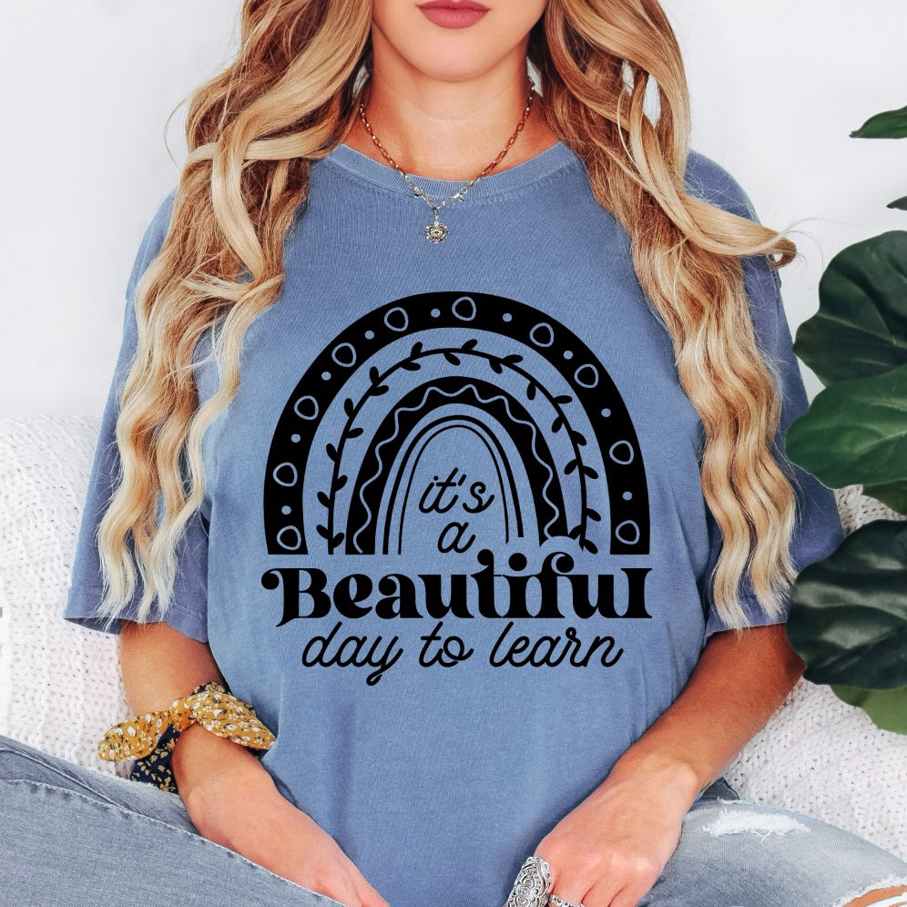 A woman with long, wavy blonde hair sits cross-legged on a light surface, surrounded by plants. She wears the Beautiful Day to Learn • Comfort Colors Tee, a blue garment-dyed shirt featuring Its a Beautiful day to learn and a black rainbow graphic.