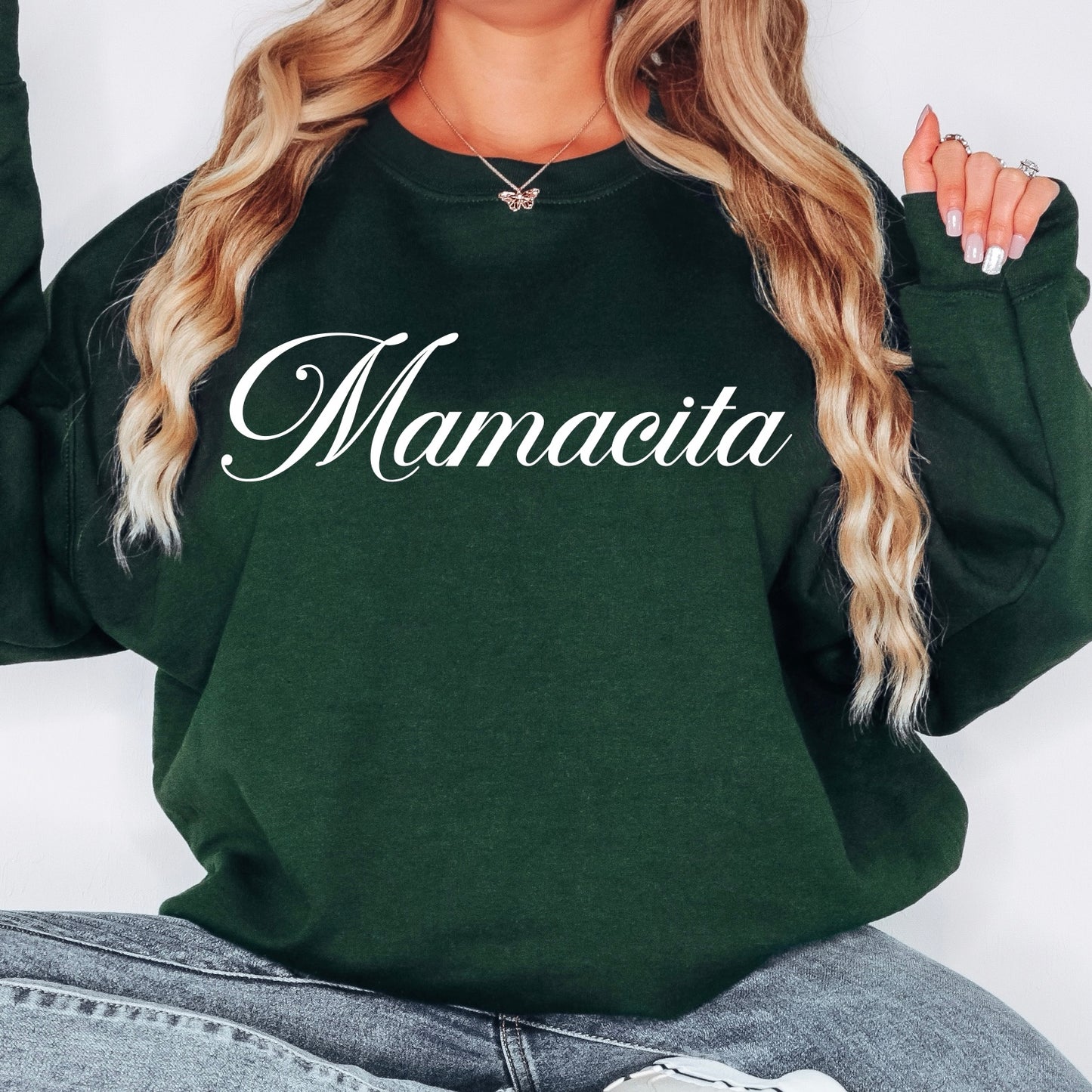 A person with long, wavy blonde hair wears the New Mamacita • Forest Pullover, a dark green unisex sweatshirt made from a soft cotton/polyester blend and featuring Mamacita in elegant white script. Only their lower face and torso are visible.