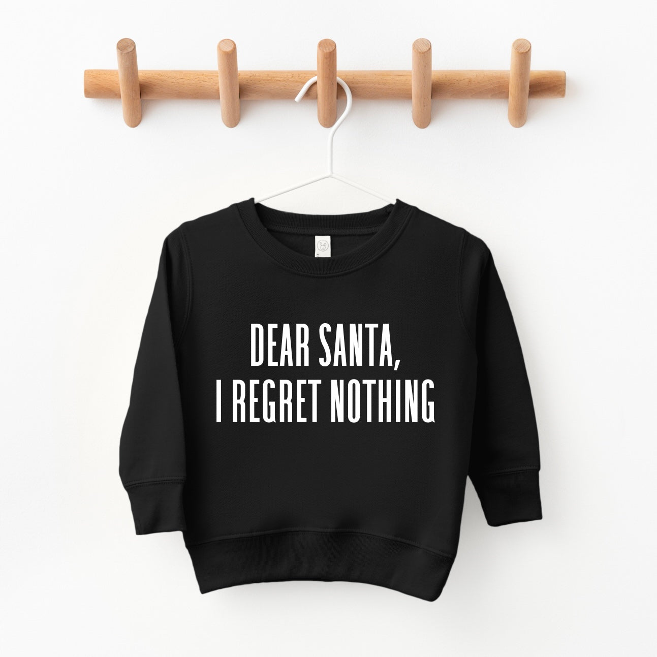 The Dear Santa • Kids Pullover, a black unisex sweatshirt with bold white DEAR SANTA, I REGRET NOTHING text, hangs on a wooden peg rack. Available in toddler/youth sizes—perfect for shopping small this season.