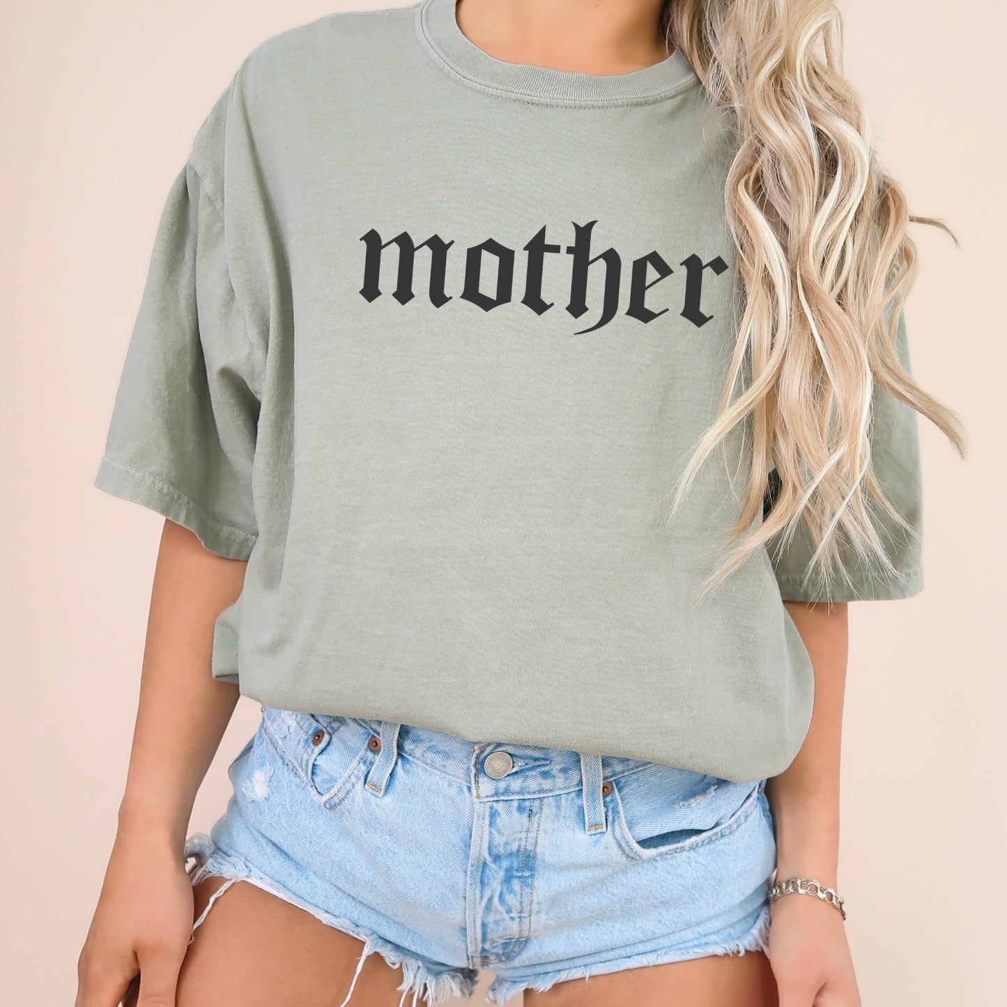 A person with long, wavy blonde hair wears the Mother • Comfort Colors Tee in light green with mother in black gothic font, styled with light blue denim shorts. The garment-dyed shirt pops against a neutral backdrop.