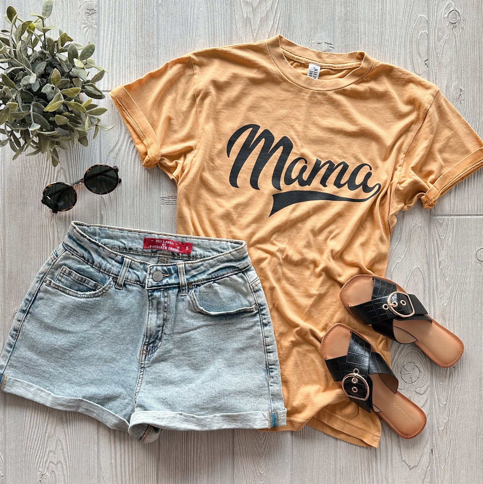 Flat lay of a casual outfit featuring the Mama Sporty • Rocker Tee in mustard burnout, light denim shorts, black slide sandals, sunglasses, and a small green plant on a light wooden surface.