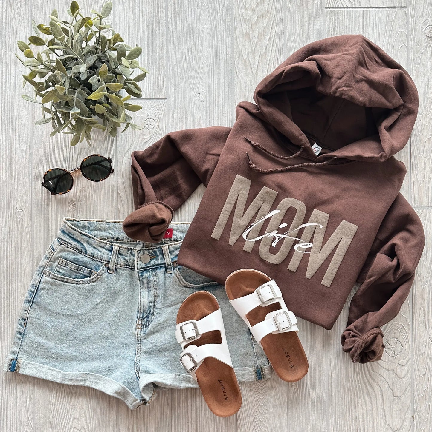 Flat lay of the Mom Tonal • Chocolate Hoodie with MOM life print, paired with light denim shorts, white sandals, black sunglasses, and a small potted plant—this cozy unisex fleece channels Pantone 2025’s trending style on a light wood background.