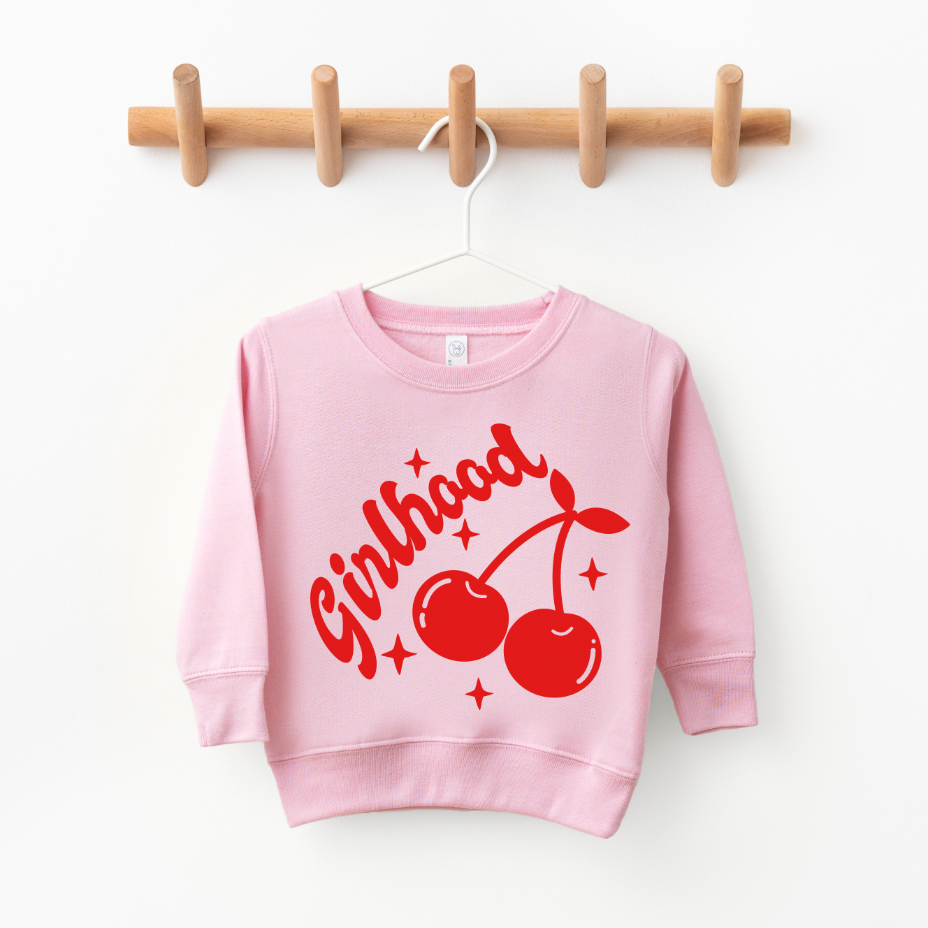 The Girlhood π β’ Pink Pulloverβ a pink childrenβs sweatshirt with Girlhood in red, cherries, and small starsβhangs on a white hanger from a wooden rack. Perfect for Mommy & Me moments and supporting small creators.