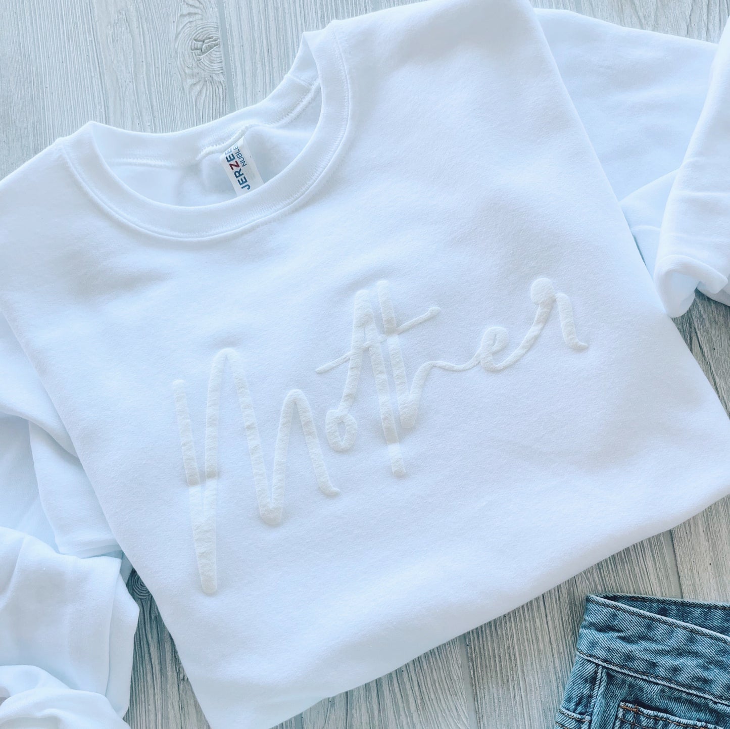 The Mother Script • Embossed White Pullover, made from a soft cotton-polyester blend with embossed lettering, is displayed on a light wooden surface next to blue denim jeans.