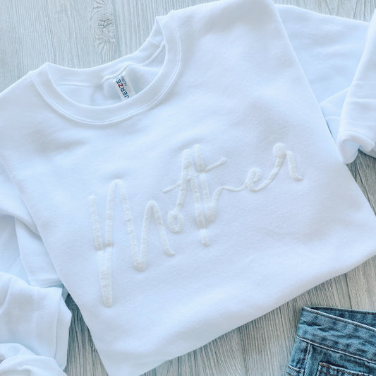 The Mother Script • Embossed White Pullover, made from a soft cotton-polyester blend with embossed lettering, is displayed on a light wooden surface next to blue denim jeans.