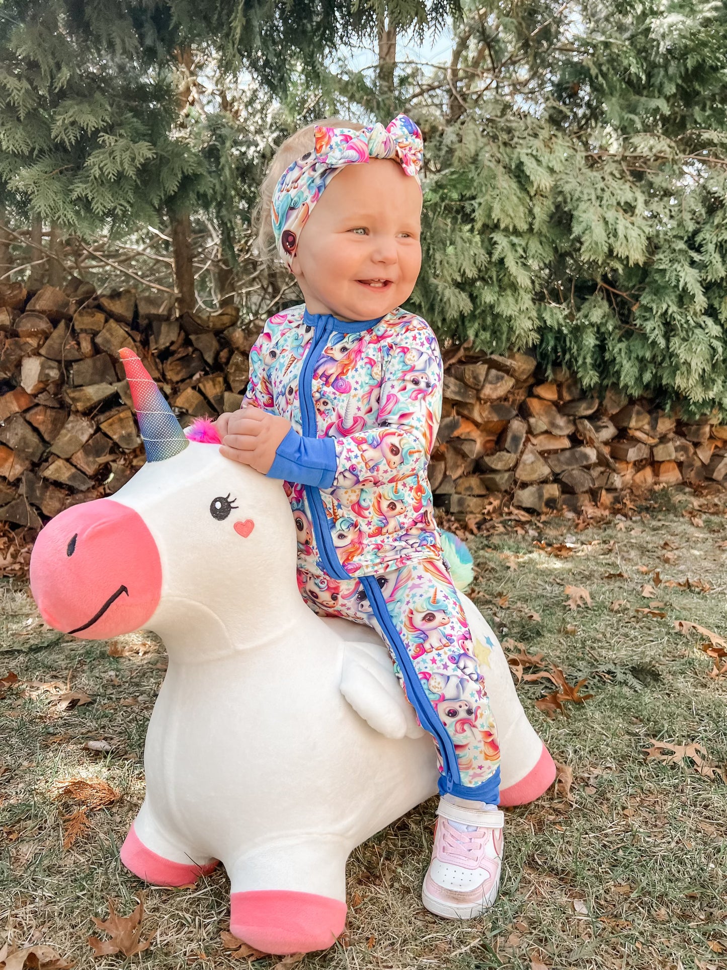 A smiling toddler in the Mythical Unicorns • Bamboo Zippy (Updated Fit) onesie sits on a white unicorn toy outdoors—colorful bamboo fabric perfect for sensitive skin.