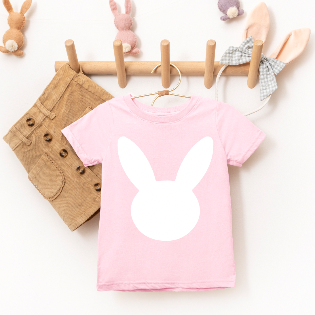 The NEW! Bunny • Light Pink Tee—a super-soft, unisex children’s t-shirt featuring a large white bunny silhouette—hangs on a peg rack beside tan shorts, with plush bunny decorations and a checkered bow in the background.