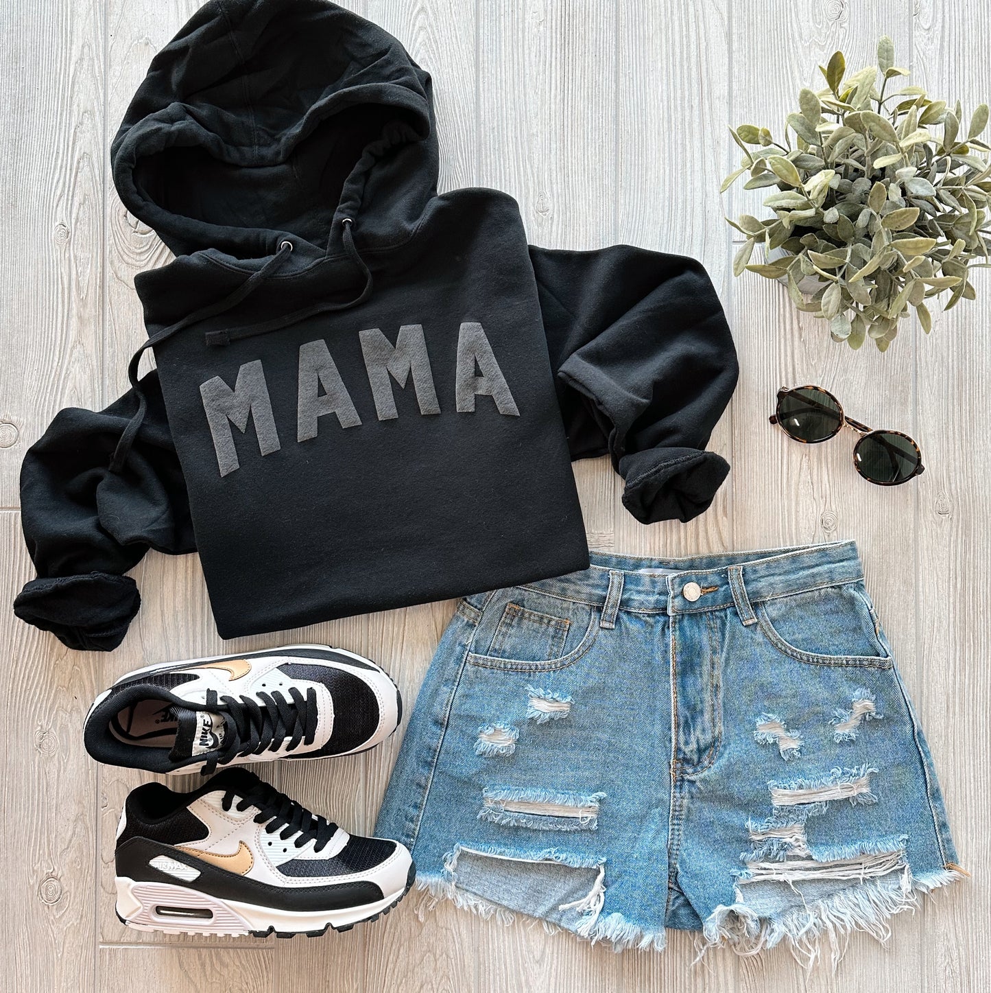 Flat lay of the MAMA • Black Tonal Hoodie with distressed denim shorts, black and white sneakers, round sunglasses, and a small potted plant on a light wooden surface. Unisex fit for everyday comfort.