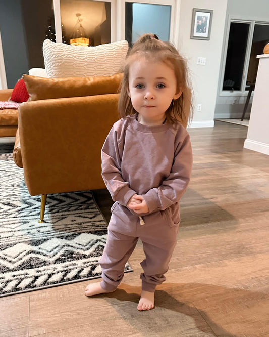 A young child with light brown hair in a ponytail stands barefoot on a wooden floor, wearing the Lilac • Sweatpant & Crew Set. A brown sofa and patterned rug are visible in the background.