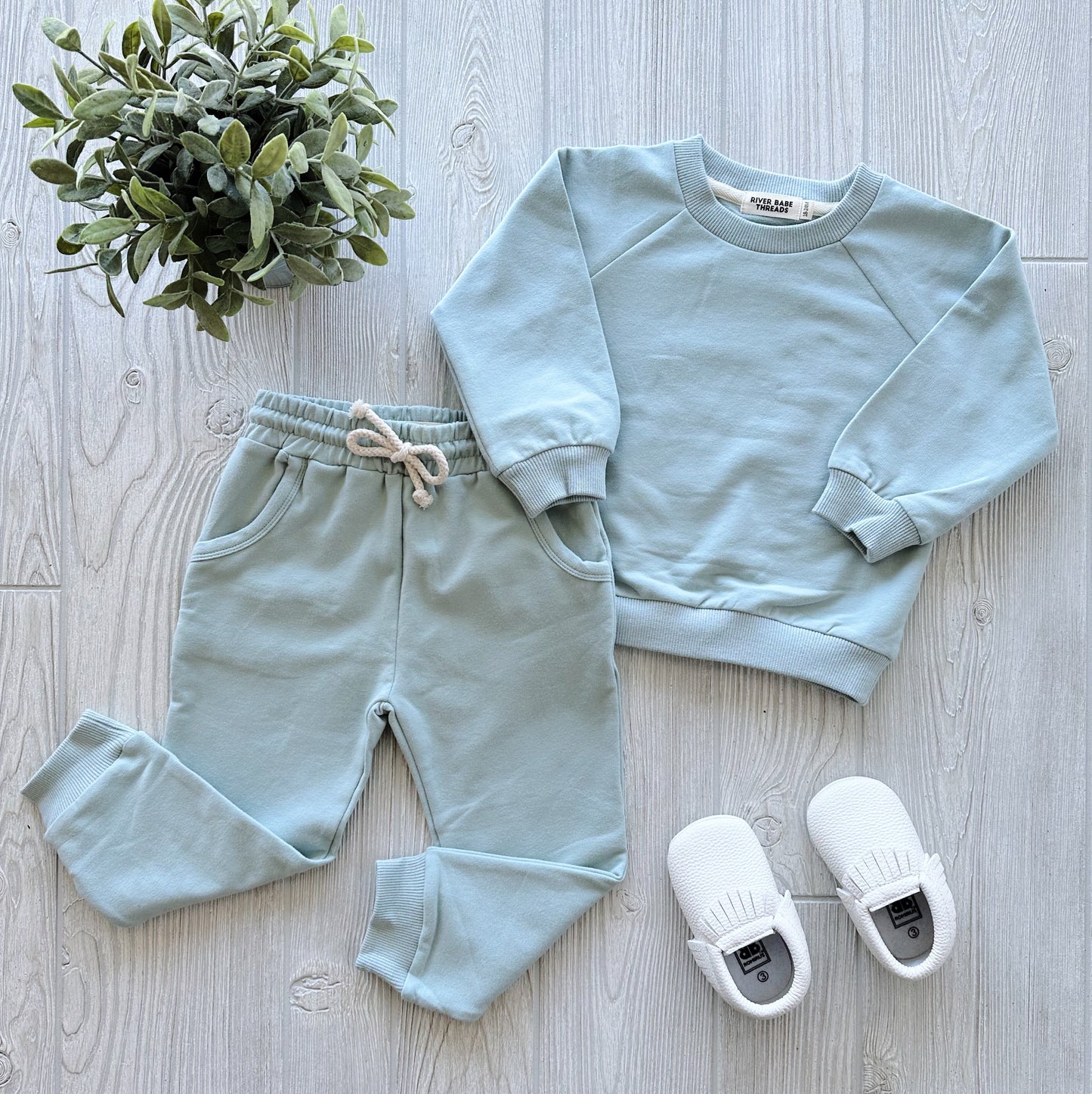 The Mint • Sweatpant & Crew Set in light blue is displayed with white slip-on shoes on a light wooden floor, next to a small potted green plant.