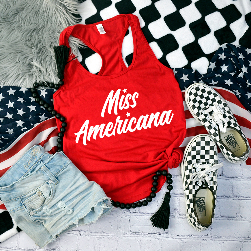 The Miss Americana • Red Tank, a soft cotton/poly blend racerback with white cursive text, is styled with denim shorts, checkered Vans, a black bead necklace, and American flag accessories on a brick backdrop.