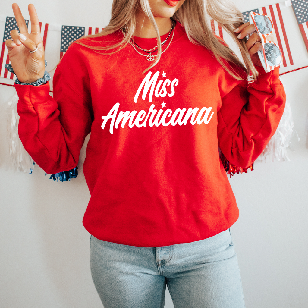 A woman wears the Miss Americana • Red Pullover with blue jeans and sunglasses, flashing a peace sign. American flag bunting in the background enhances the fun, patriotic atmosphere.