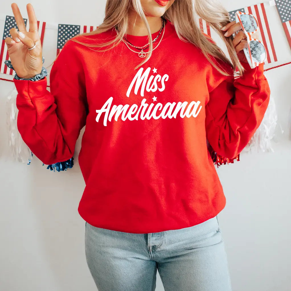 A woman wears the Miss Americana • Red Pullover with blue jeans and sunglasses, flashing a peace sign. American flag bunting in the background enhances the fun, patriotic atmosphere.