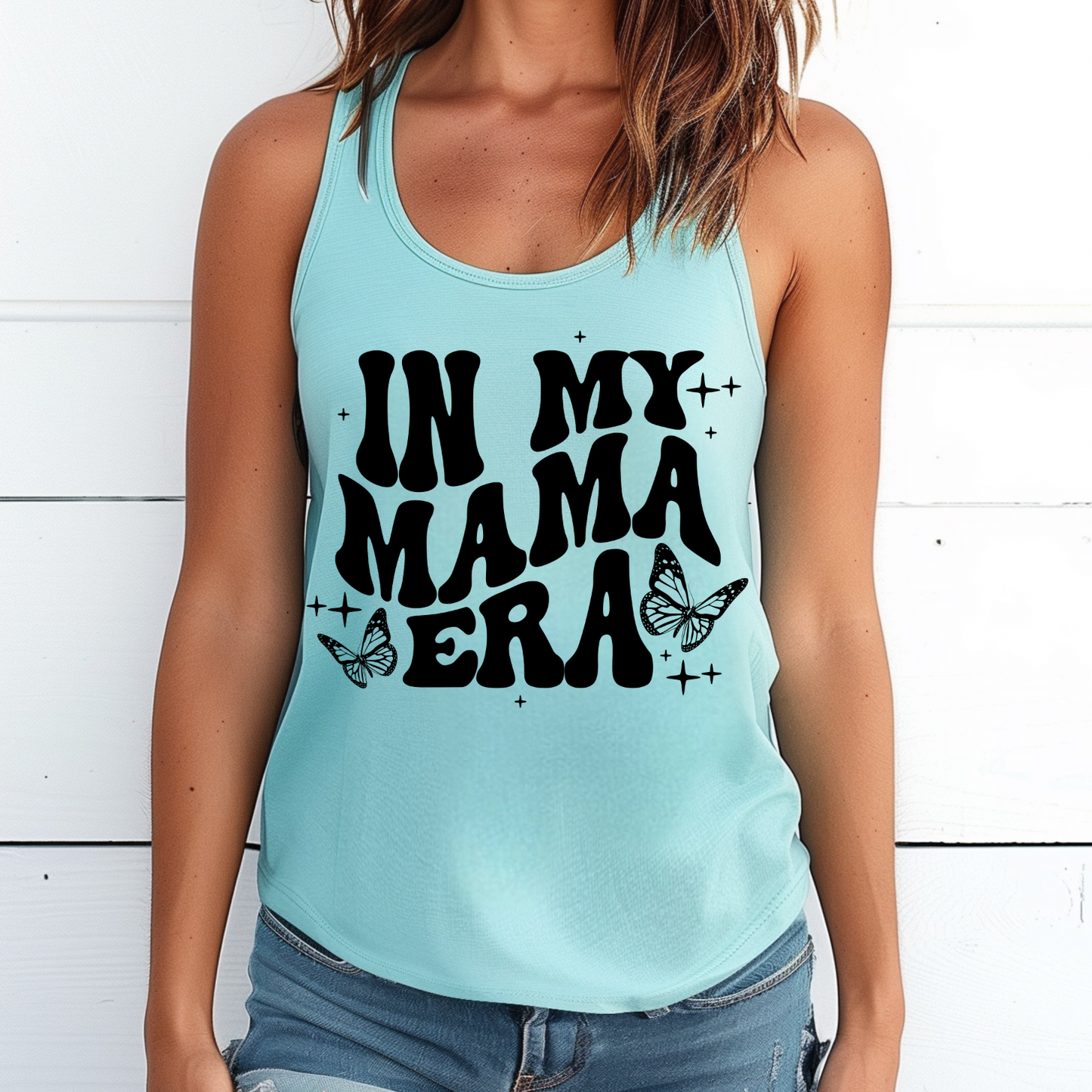 A woman wears the Mama Era • Cancun Tank, a light blue racerback with bold black IN MY MAMA ERA text and butterfly designs. Standing by a white wooden wall, this tank is a perfect addition to your summer closet.