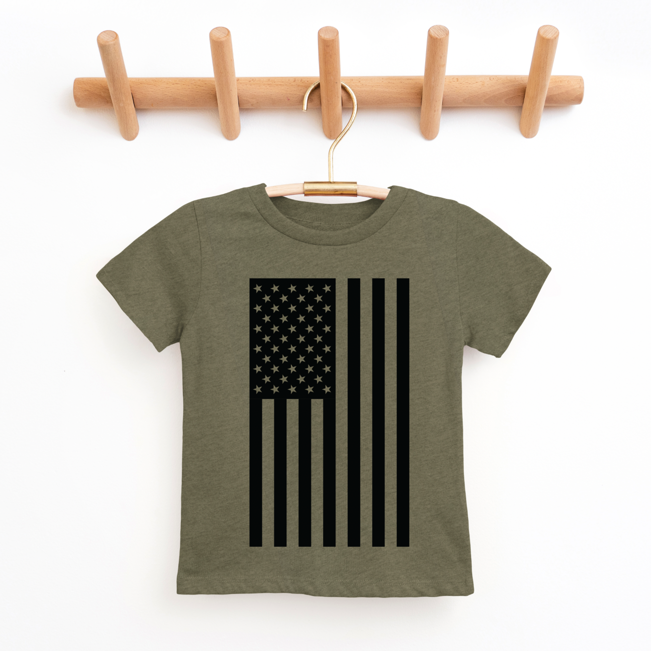 The Flag • Kids Tee by Bella/Canvas, a green unisex children’s t-shirt with a black vertical American flag design, hangs on a wooden hook rack against a white background.