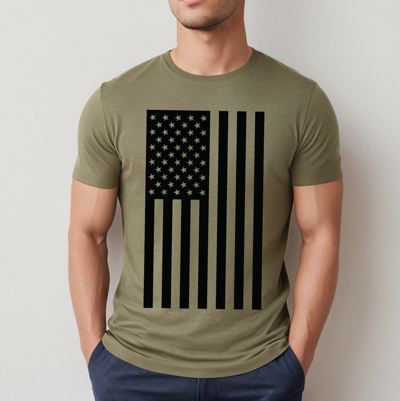Someone wearing the Flag Tee • Adult in olive green, which features a large black American flag with vertical stripes and stars, stands against a plain light wall with hands in pockets.