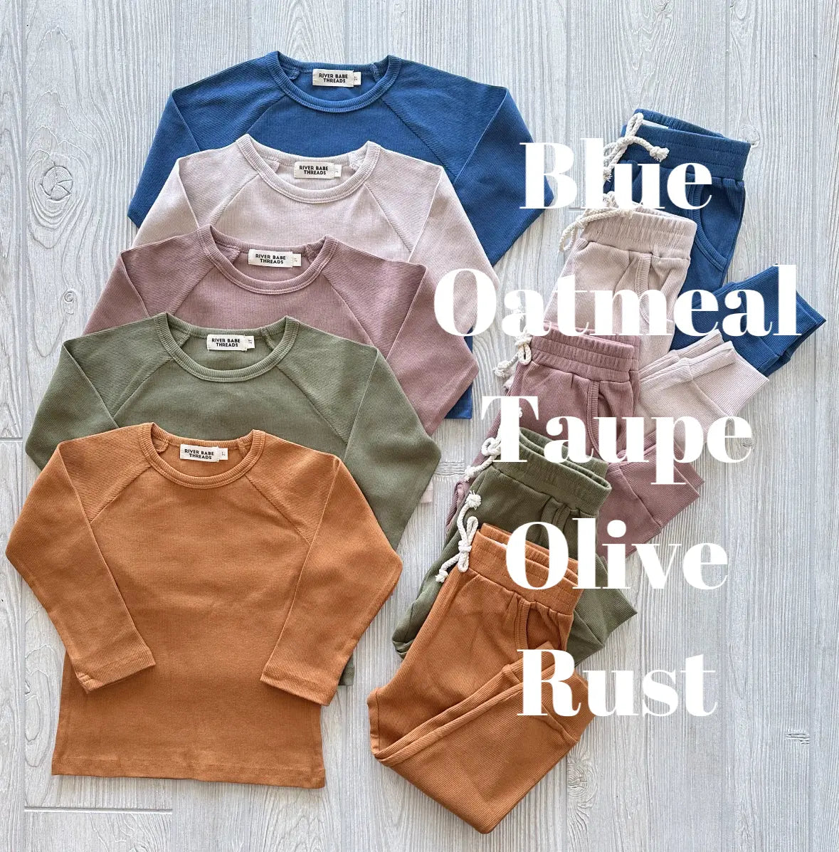 Five Ribbed Organic Cotton Jogger Sets—long-sleeve shirts and pants in blue, oatmeal, taupe, olive, and rust—are neatly arranged on a light wood floor. Each sets color is labeled in large white text.