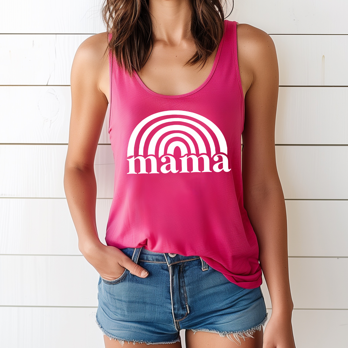 A woman wears the Mama Rainbow • Raspberry Tank, a vibrant pink racerback with mama and a rainbow graphic in a flattering womens fit, standing by a white wooden wall with one hand in her denim shorts pocket.