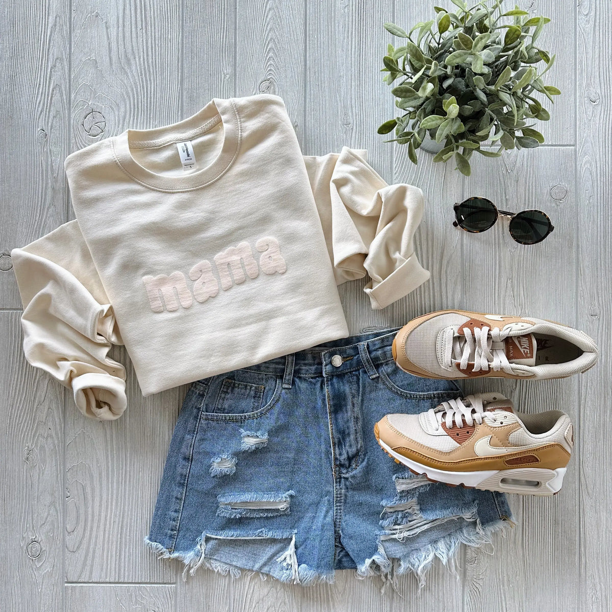 The Mama Wave • Marshmallow Tonal unisex crew neck sweatshirt is styled with distressed denim shorts, beige and white Nike sneakers, round sunglasses, and a small potted plant on a light wooden floor.