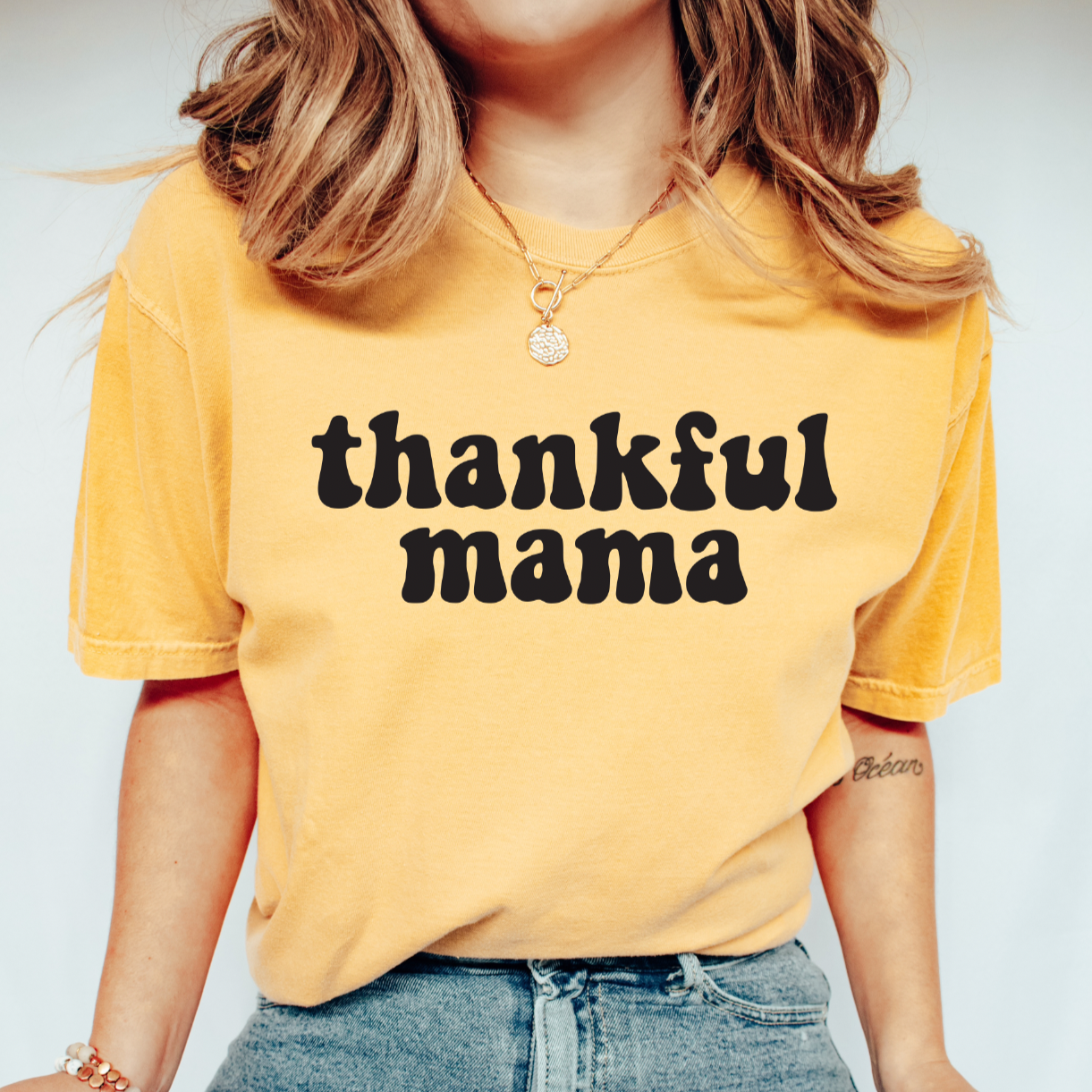 A woman wears the thankful mama • Mustard Tee from the vintage collection, featuring bold black lettering. She pairs it with blue jeans, a gold necklace, and has wavy hair for a classic style.
