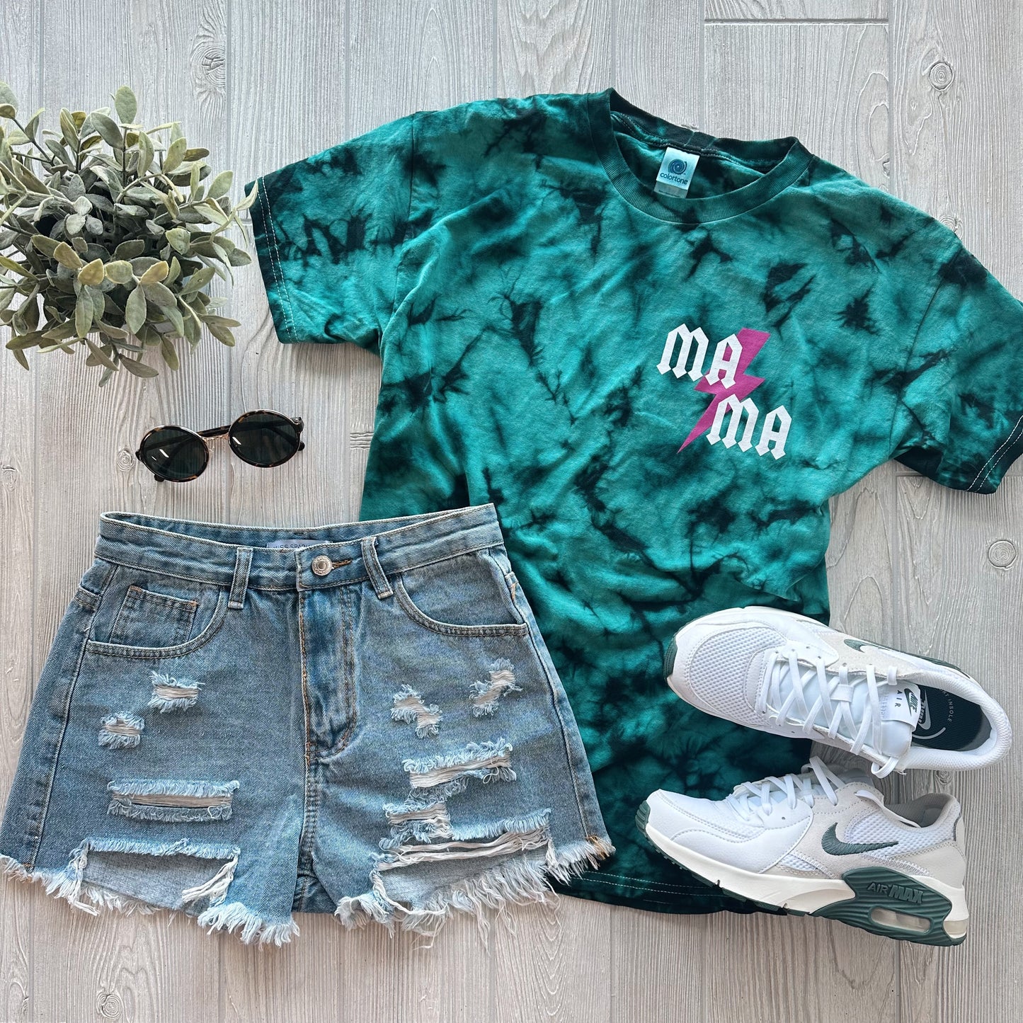 A flat lay of the MA⚡︎MA Left Chest • Teal Tie-Dye unisex tee paired with ripped denim shorts, white sneakers, black sunglasses, and a small potted plant on a light wood floor.