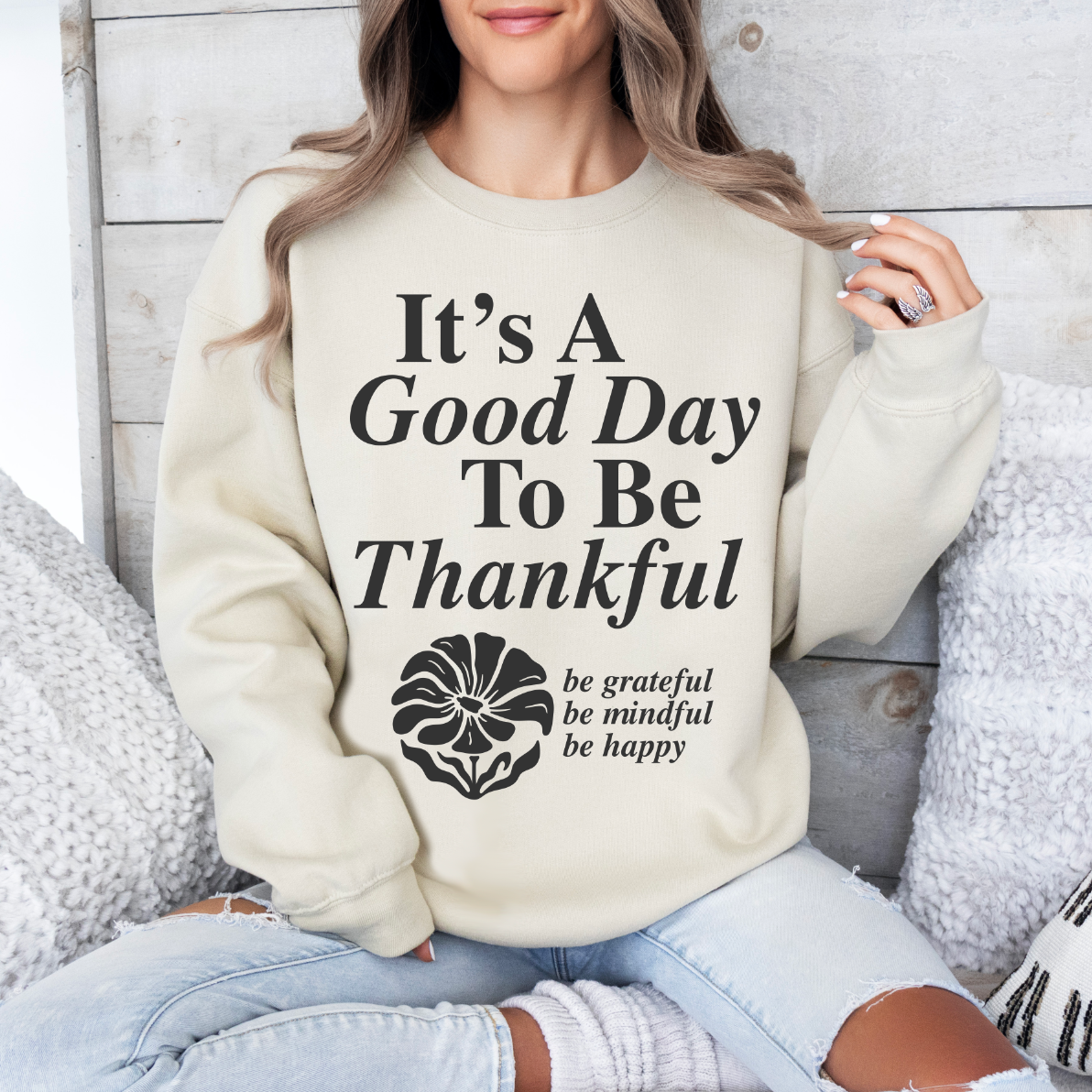 A woman relaxes on a couch, smiling in the Good Day to be Thankful • Sand Pullover—a cozy unisex sweatshirt perfect for enjoying cooler weather and a grateful mindset.