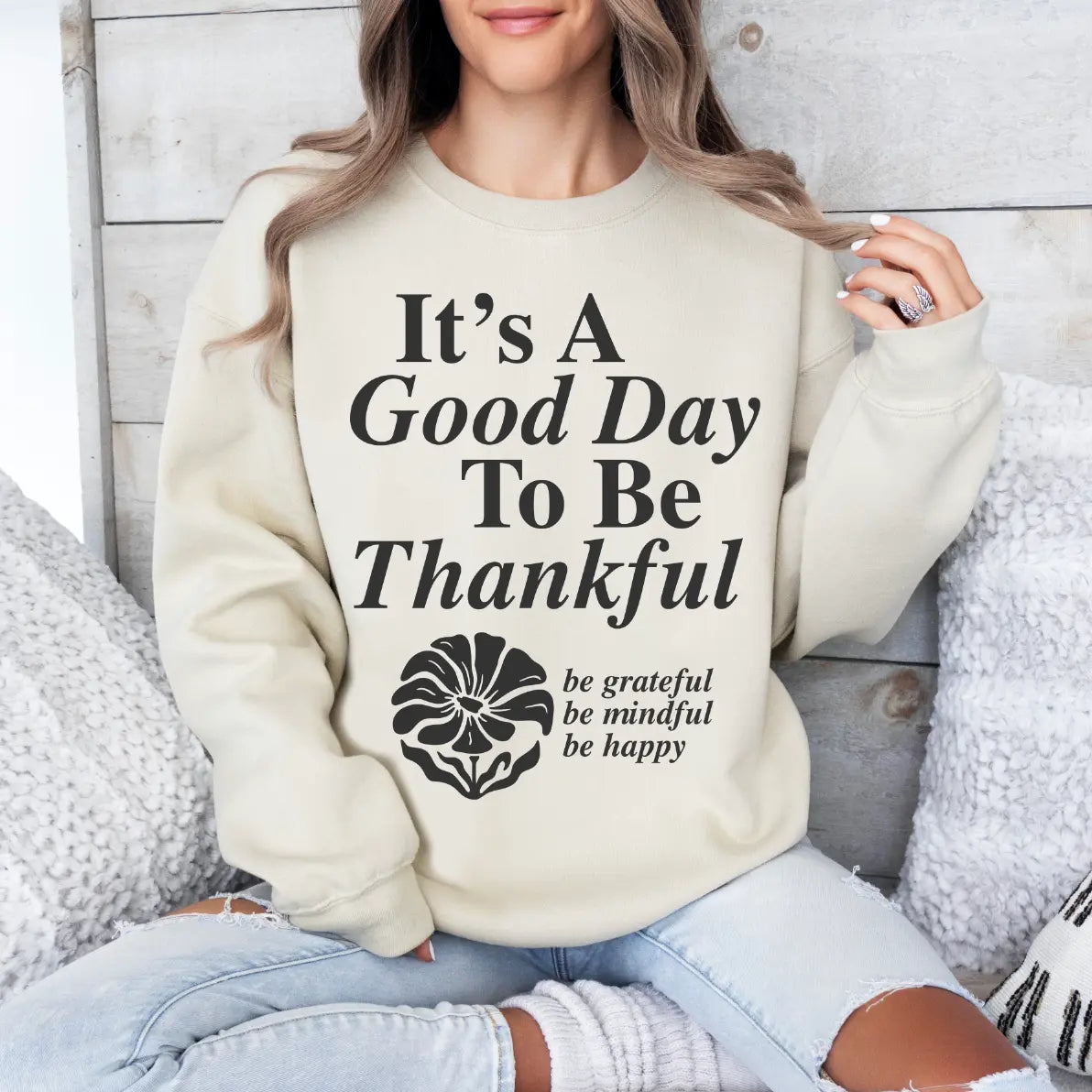 A woman relaxes on a couch, smiling in the Good Day to be Thankful • Sand Pullover—a cozy unisex sweatshirt perfect for enjoying cooler weather and a grateful mindset.