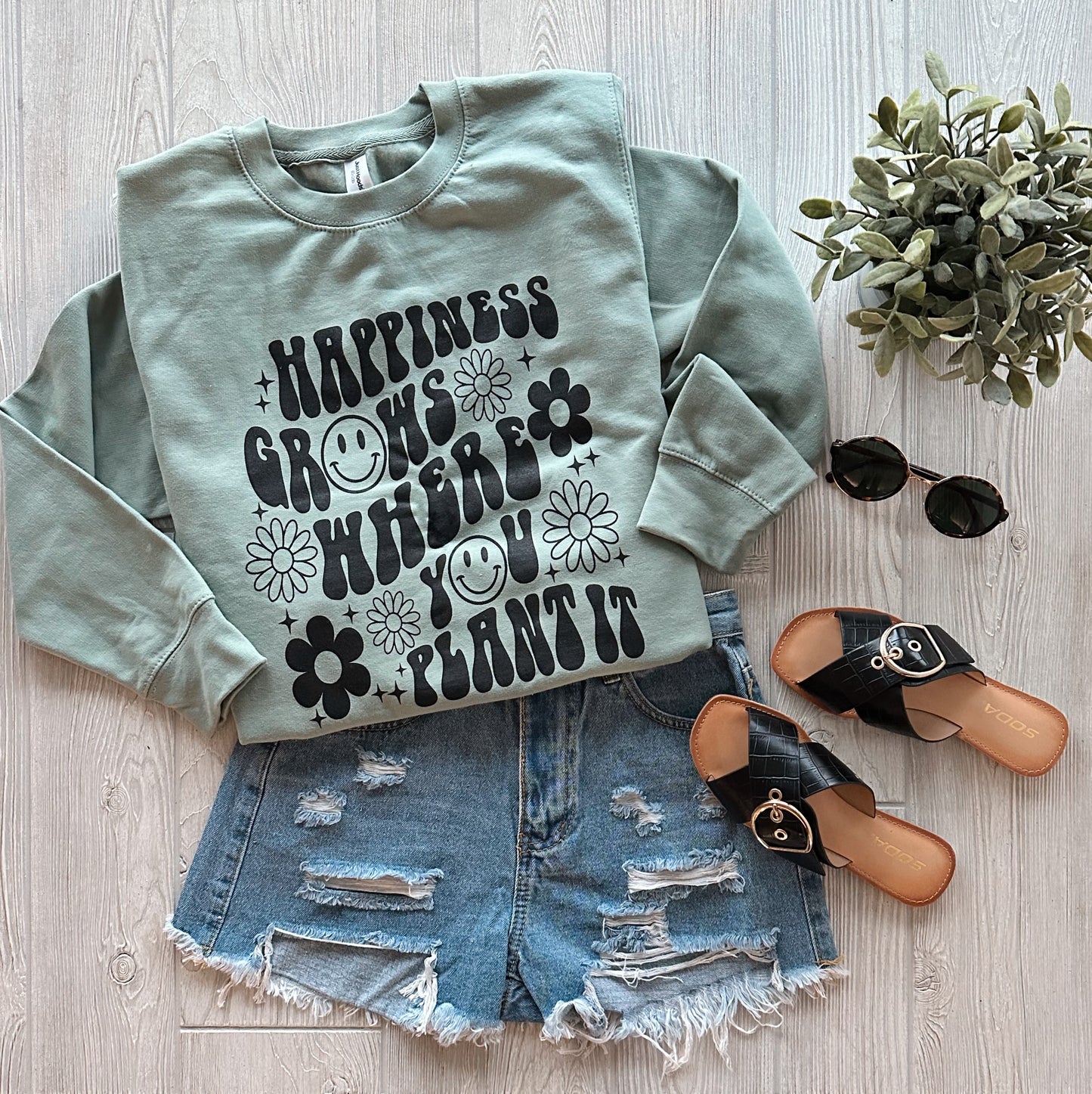 The HAPPINESS • Sage Pullover, a sage green cotton-polyester blend with Happiness Grows Where You Plant It in black, is styled with distressed denim shorts, black sandals, sunglasses, and a potted plant on a light wood floor.