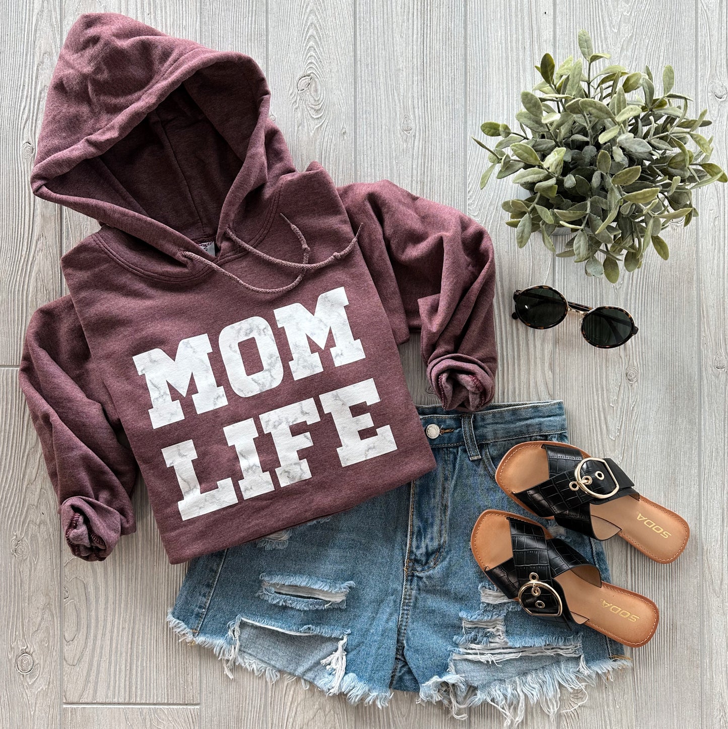A flat lay of a maroon MOM LIFE Marble • Maroon Hoodie with a unisex fit, styled with distressed denim shorts, black sandals, round sunglasses, and a small potted plant on a light wooden floor.