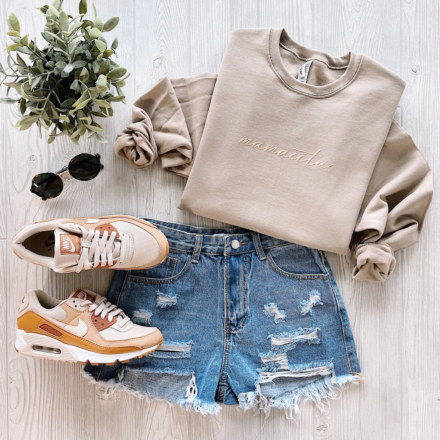 Flat lay of a casual outfit featuring the mamacita • Khaki Pullover, distressed denim shorts, tonal beige and white sneakers, round sunglasses, and a small green potted plant on a light wooden surface.