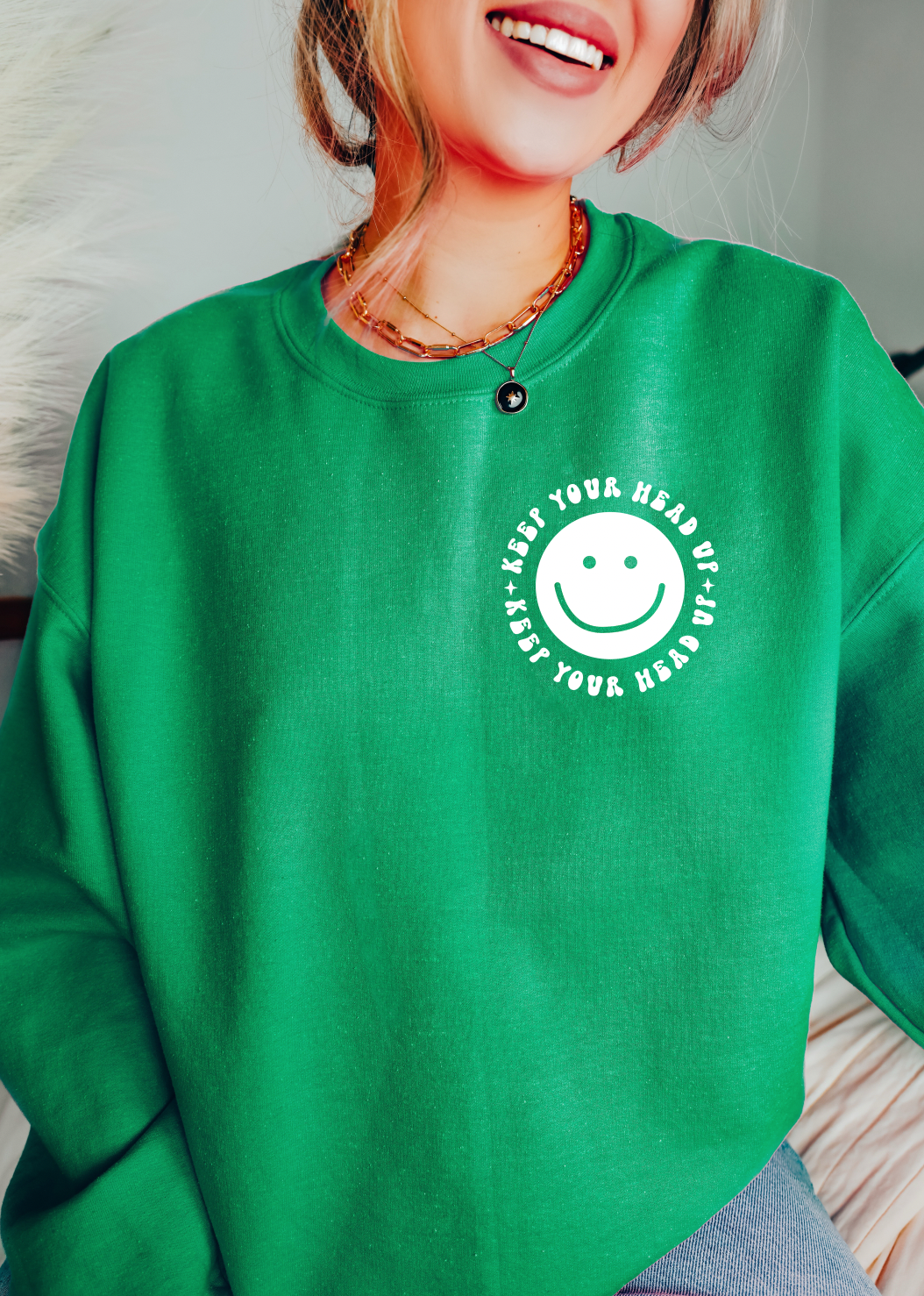 A smiling person wears the Keep Your Head Up • Mental Health Pullover, a green unisex sweatshirt with a smiley face and uplifting “KEEP YOUR HEAD UP!” text—designed to inspire positivity every day.