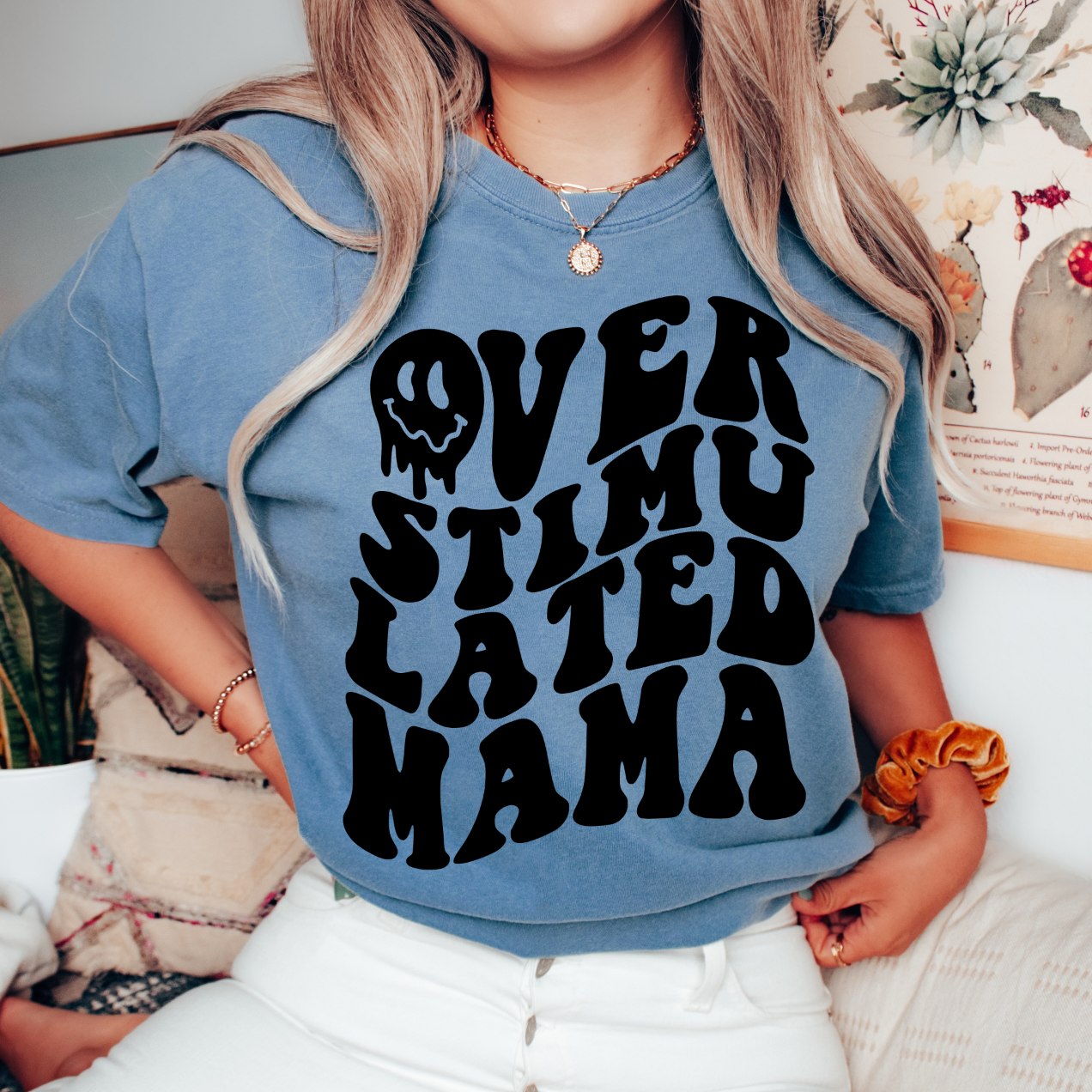 A woman wears the Overstimulated • Blue Jean Dyed Tee, featuring bold wavy black text. This comfy unisex shirt is perfect for summer lounging, as she relaxes indoors on a couch surrounded by plants and floral art.