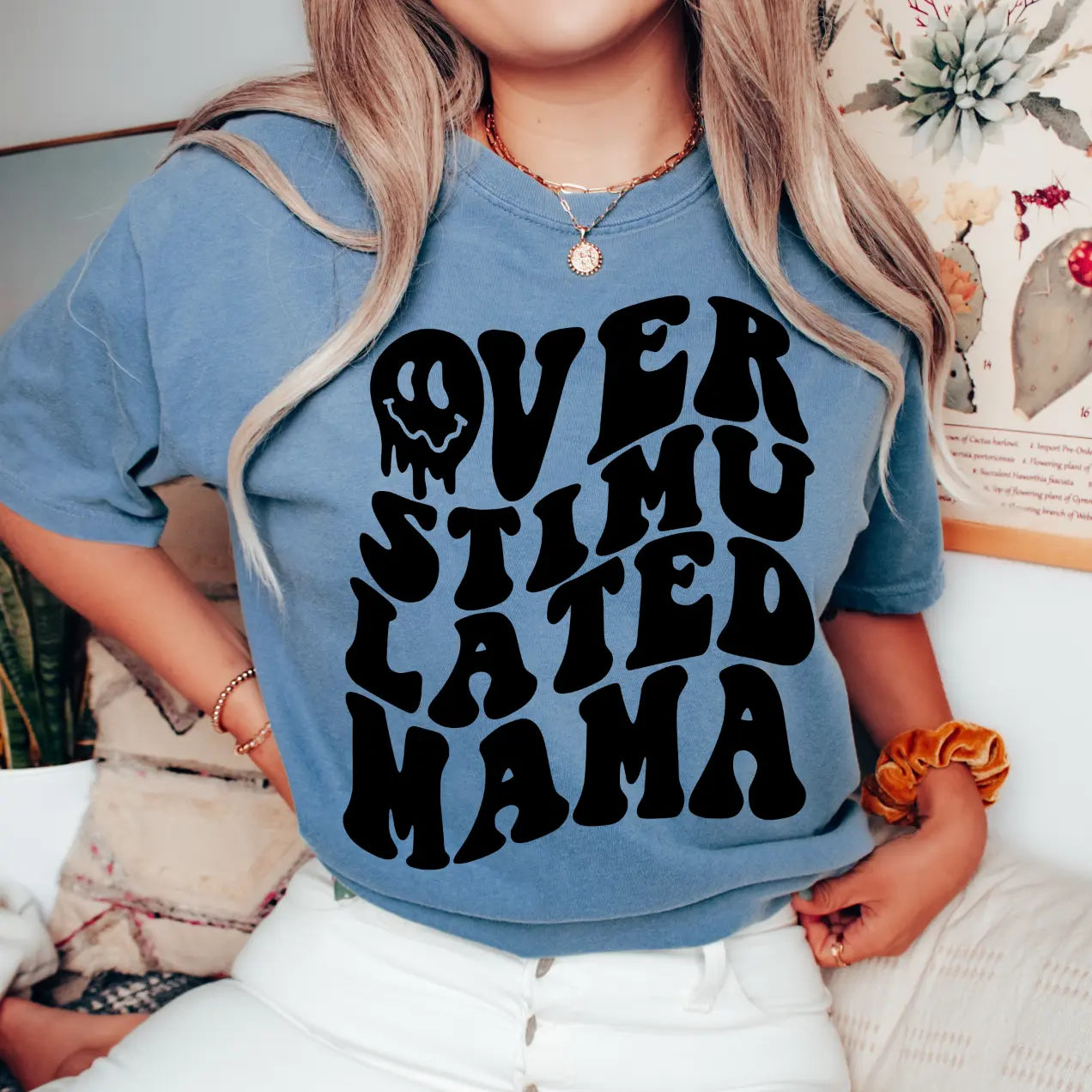 A woman wears the Overstimulated • Blue Jean Dyed Tee, featuring bold wavy black text. This comfy unisex shirt is perfect for summer lounging, as she relaxes indoors on a couch surrounded by plants and floral art.
