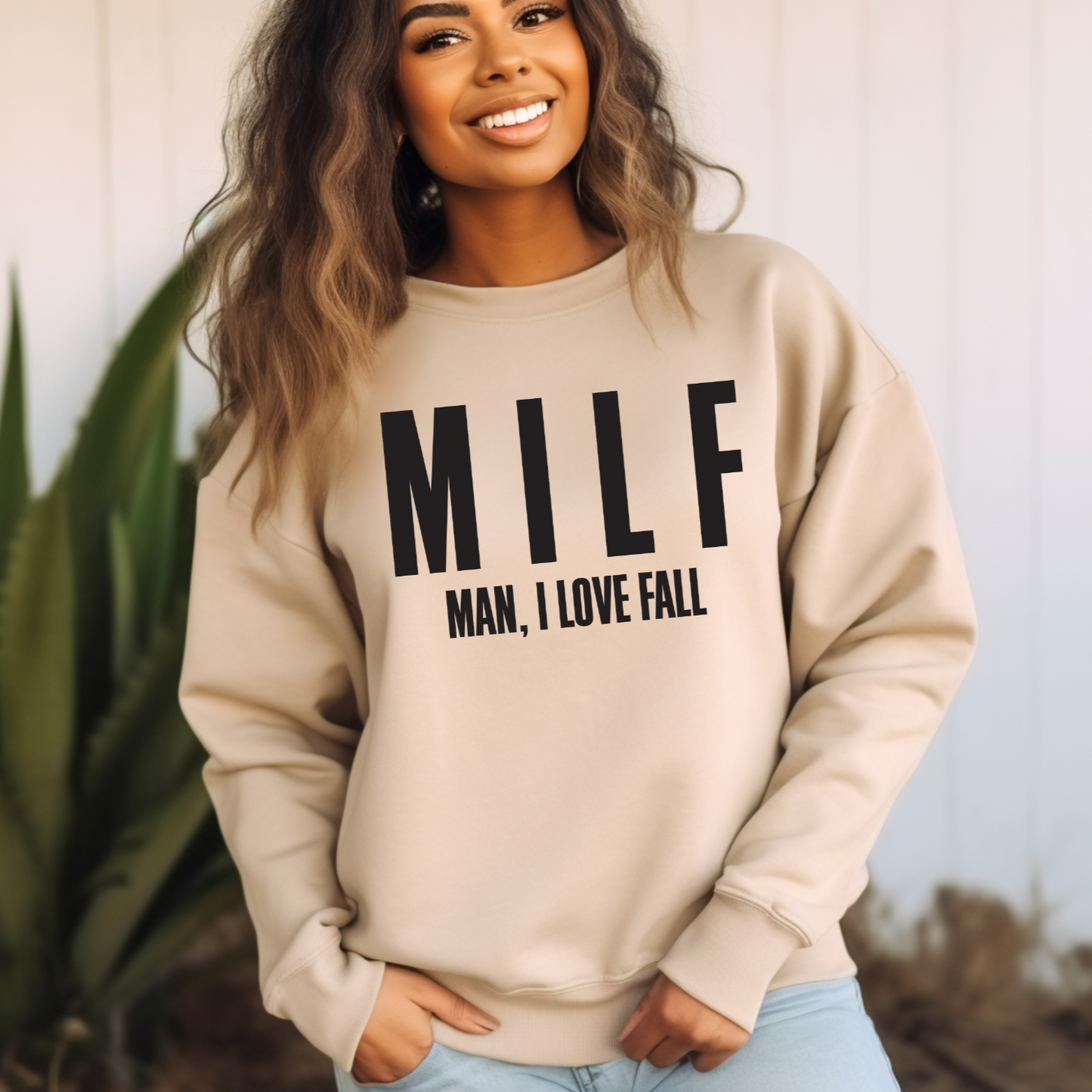 A smiling woman stands outdoors wearing the MILF • Sand Pullover featuring bold black MILF text and Man, I Love Fall underneath. Green plants and a white wall in the background highlight this cozy fall design.