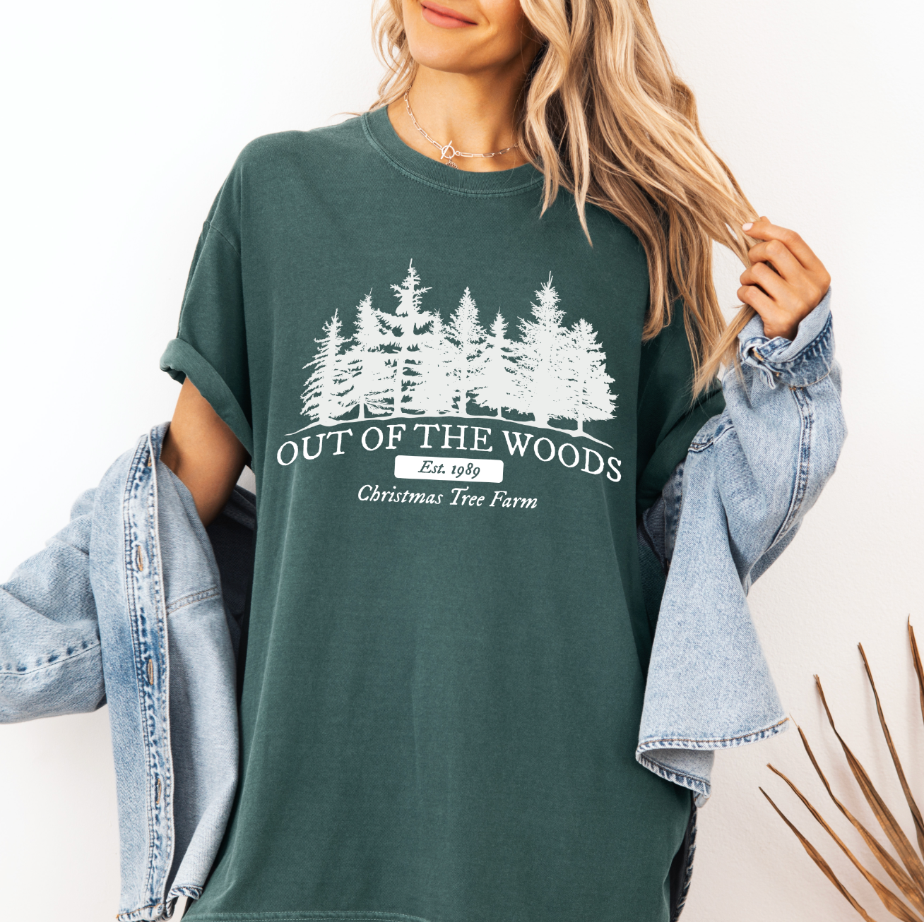A woman wears the Christmas Farm • Comfort Colors Tee in green, featuring white pine tree graphics and OUT OF THE WOODS Est. 1989 Christmas Tree Farm text, styled with a denim jacket draped over her arms.
